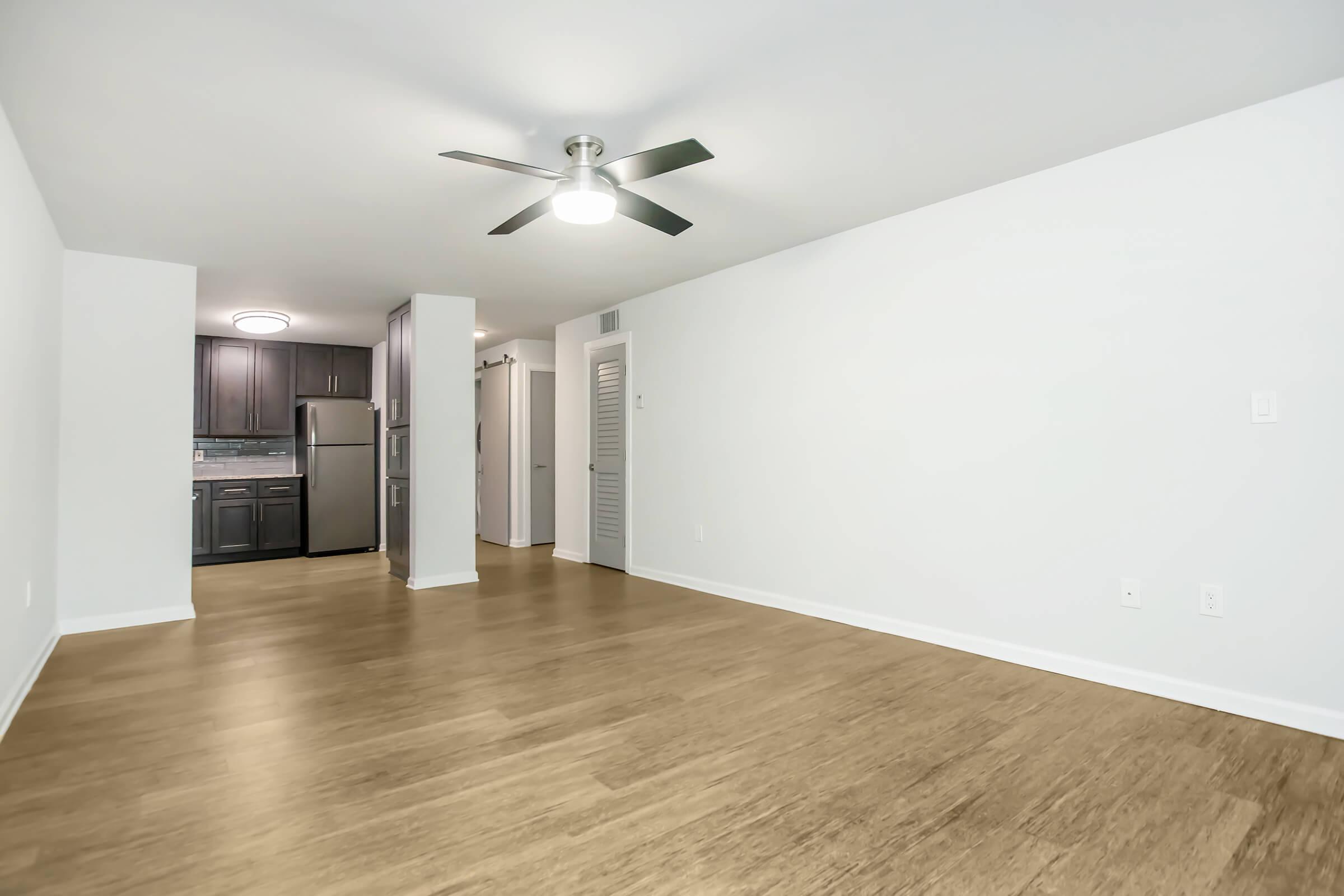 A spacious, empty living room featuring light wood flooring, white walls, and a ceiling fan. The room opens to a modern kitchen with dark cabinetry in the background, and there is a doorway leading to another area. Natural light brightens the space, creating a welcoming atmosphere.