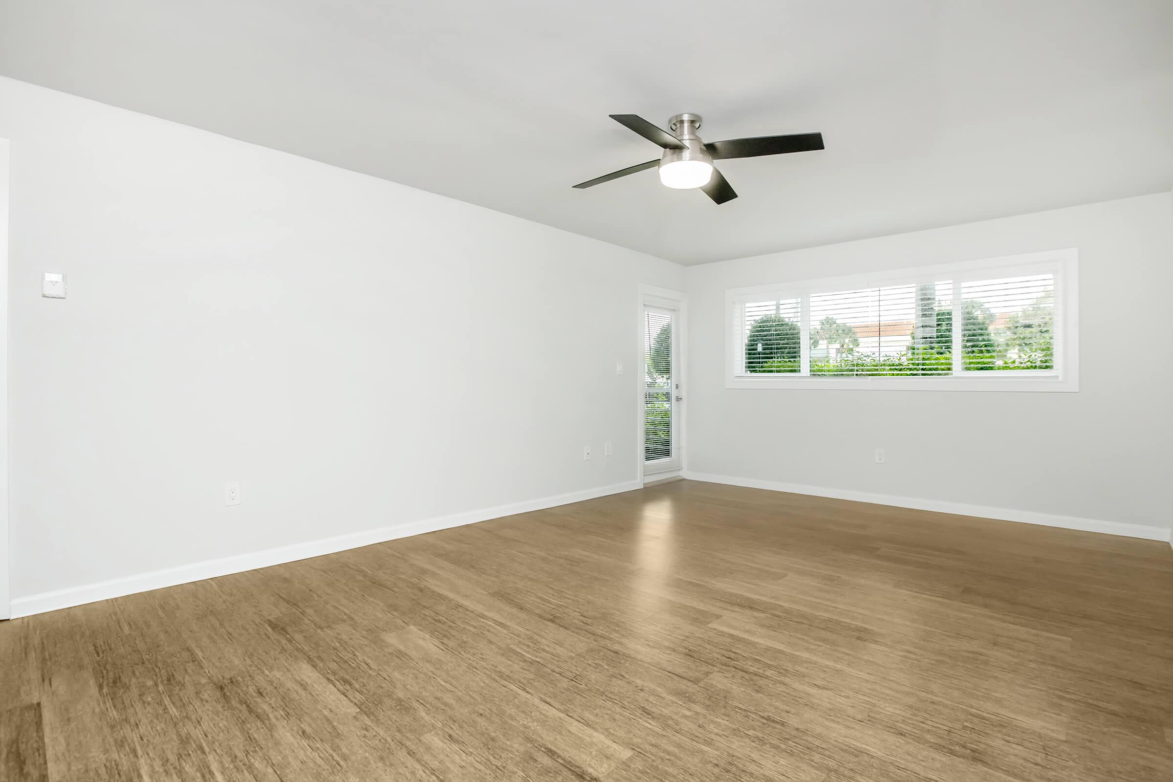Bright, spacious room with light-colored walls and a ceiling fan. Features large windows with blinds allowing natural light, and a view of greenery outside. The floor is a warm wood finish, adding a cozy touch to the modern aesthetic. Ideal for various interior design possibilities.