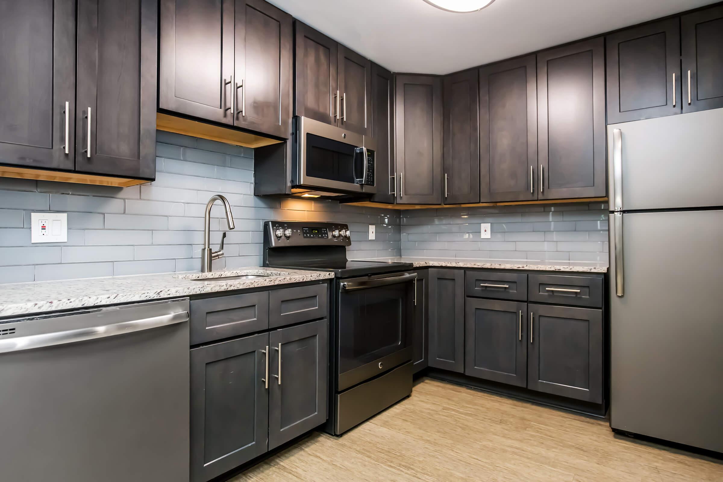 Modern kitchen featuring dark cabinetry, stainless steel appliances including a refrigerator, stove, and microwave. The countertops are made of light-colored granite, and there is a gray backsplash. The layout is compact and functional, with under-cabinet lighting and a contemporary design.