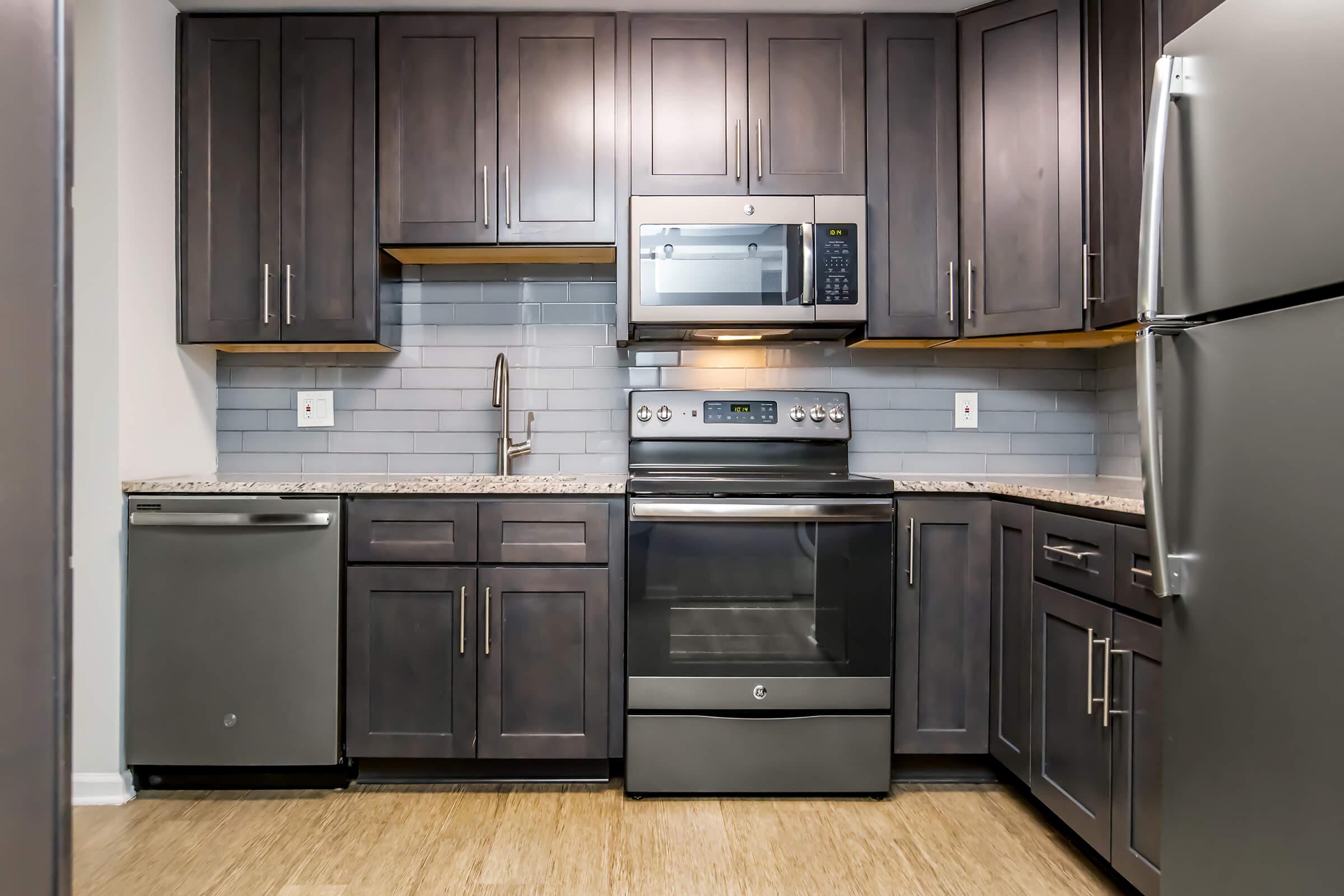 Modern kitchen featuring dark wood cabinetry, stainless steel appliances including an oven, microwave, and dishwasher, and a light-colored countertop. The backsplash consists of gray tiles, and the flooring is a warm-toned laminate. The overall design is sleek and contemporary.