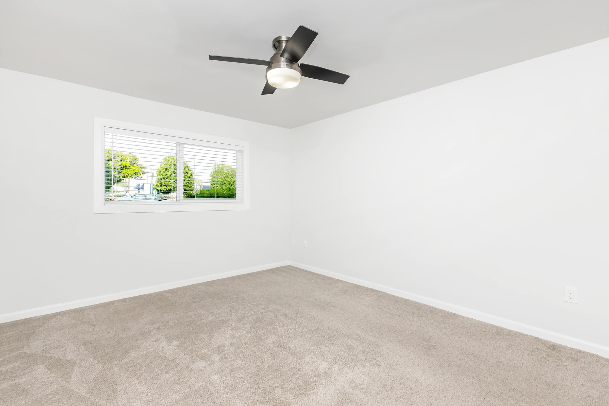 Bright, empty room with white walls and light beige carpet. A modern ceiling fan with a light is central on the ceiling. There is a window with horizontal blinds, allowing natural light and a view of greenery outside. The space is minimalistic and uncluttered, ready for furnishing.