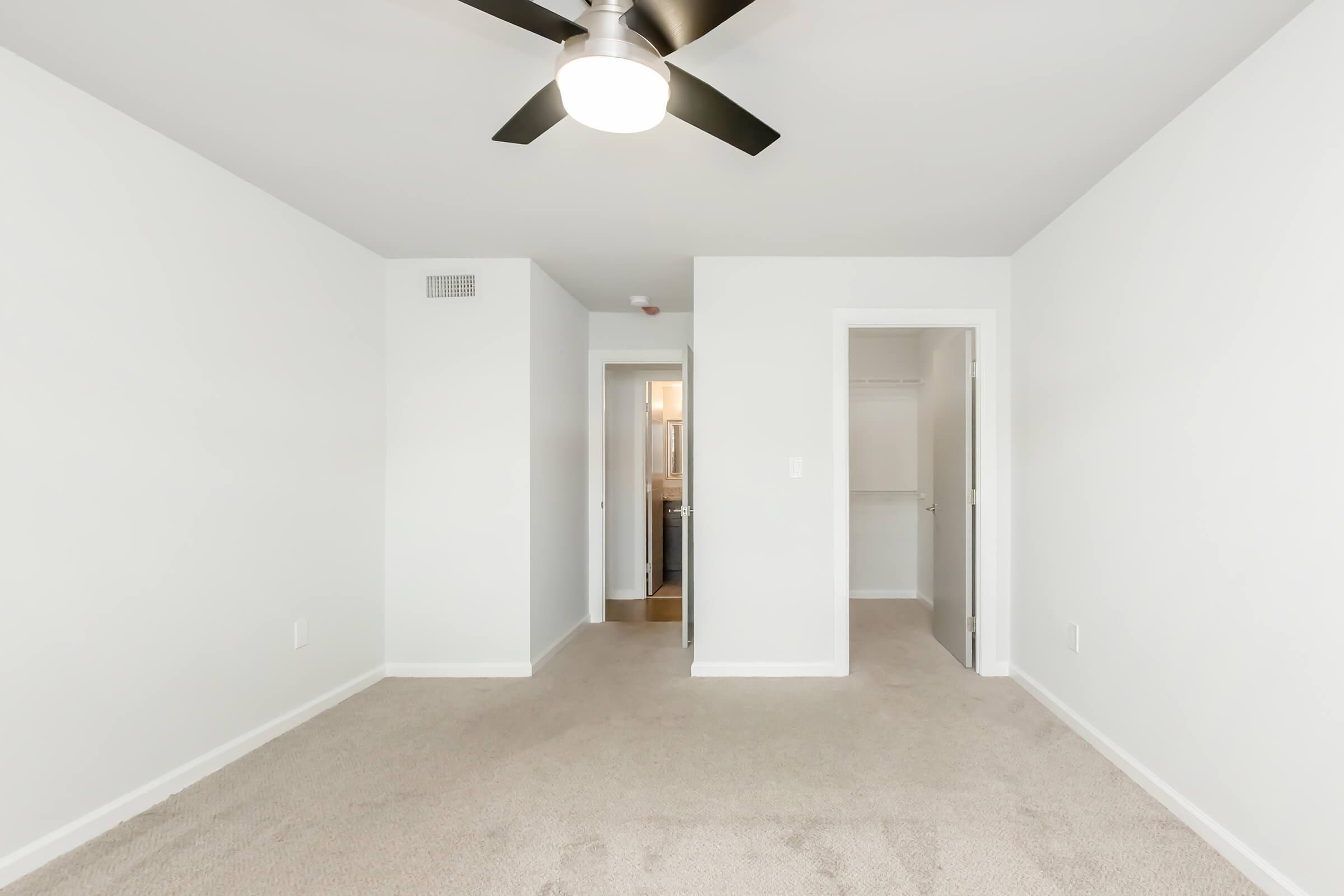 A spacious, empty bedroom with light gray walls and beige carpet. It features a ceiling fan, a door leading to a closet, and another door that opens to a bathroom. Natural light illuminates the room through the doorway. The overall atmosphere is bright and minimalist, offering a clean slate for personalization.