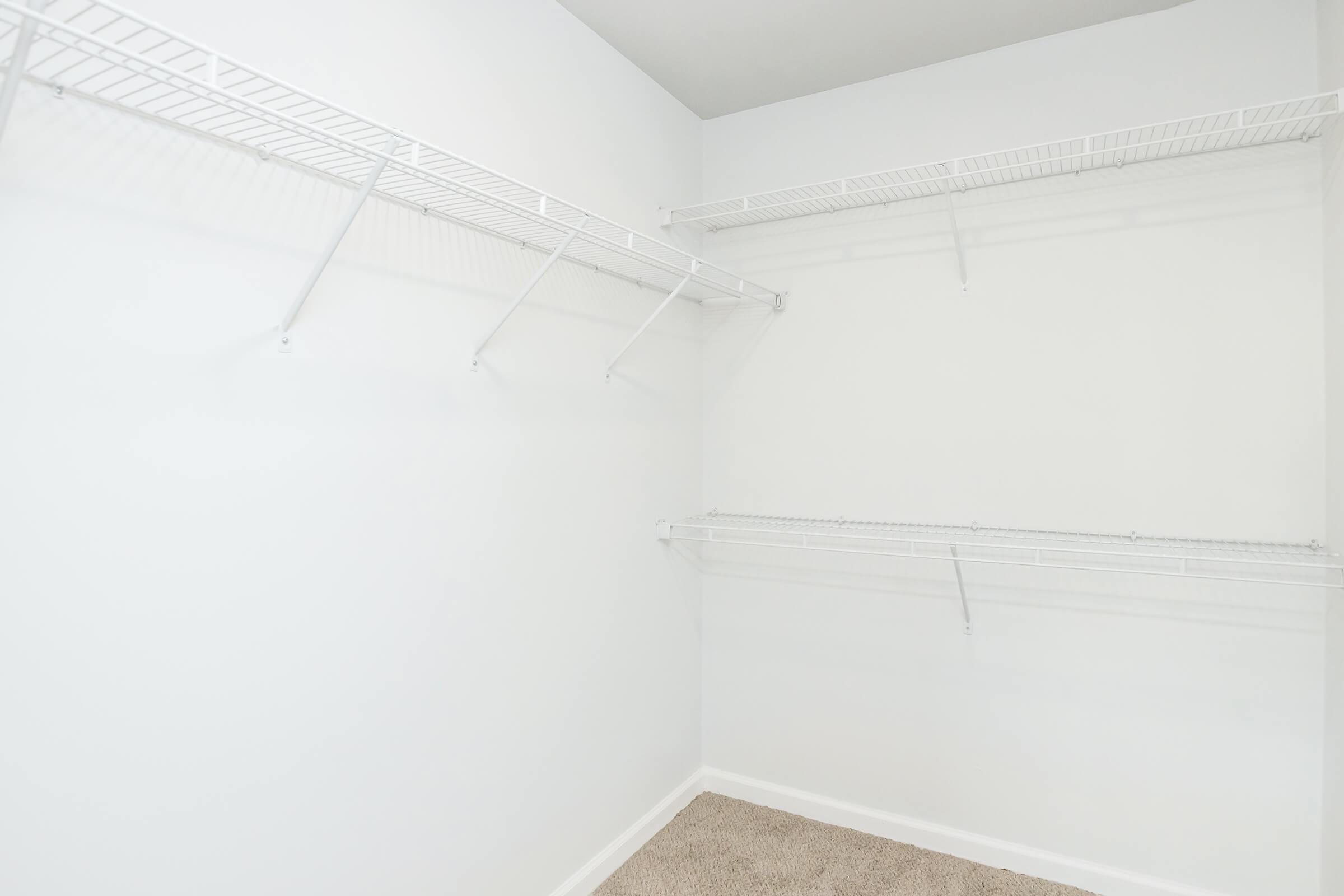 A spacious, empty closet with white walls, featuring several wire shelving units mounted on two sides. The floor is covered with light tan carpet, and the overall space is bright and uncluttered, showing potential for organization or storage.