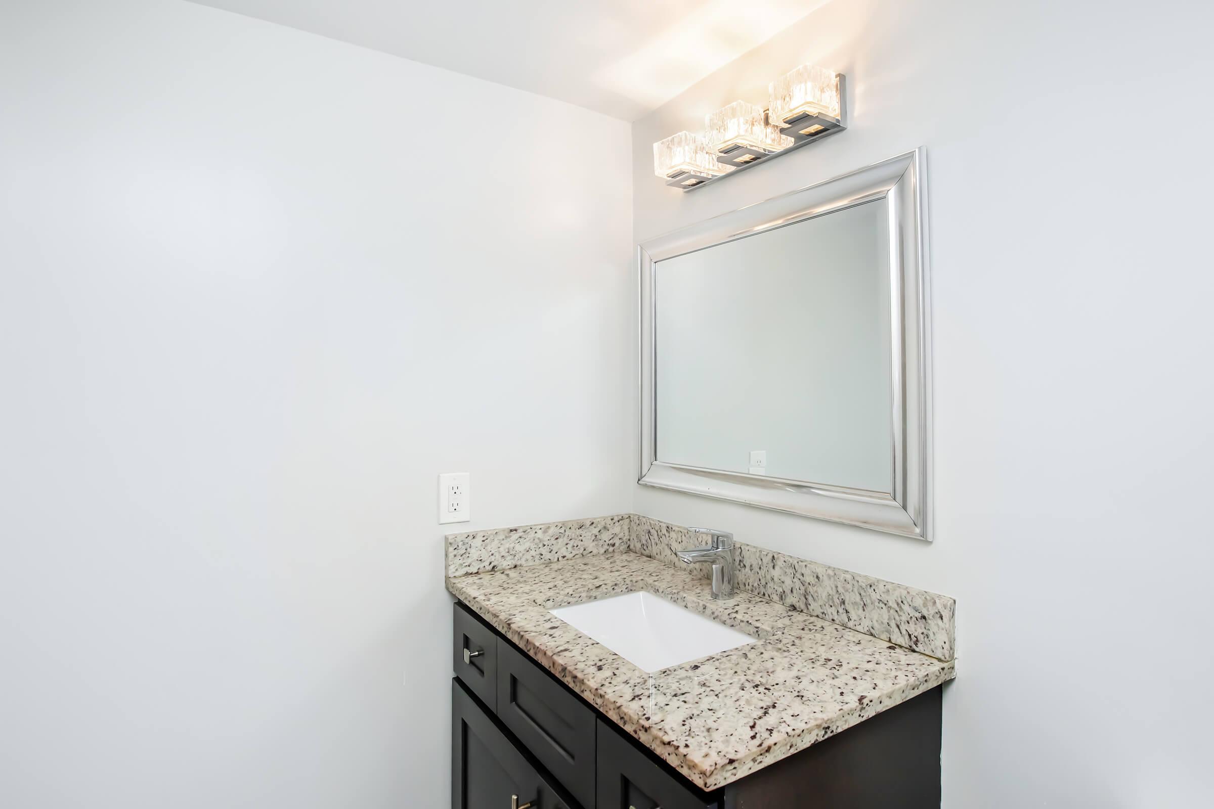 A modern bathroom vanity featuring a granite countertop with a rectangular sink, a stylish silver-framed mirror above it, and a contemporary light fixture with three glass bulbs. The wall is painted a soft white, complementing the dark cabinetry below.