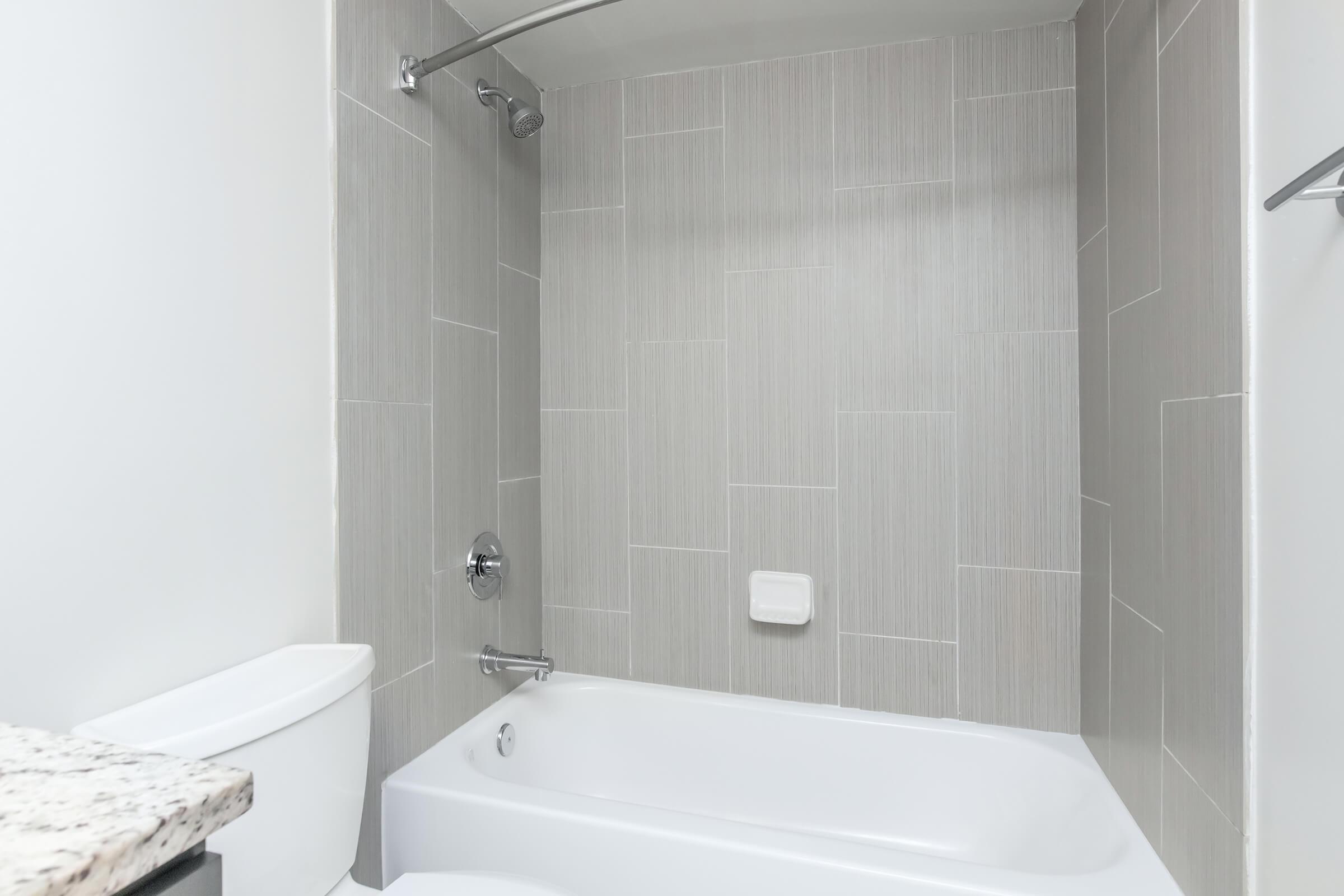 A modern bathroom featuring a white bathtub with a chrome faucet, a standing showerhead, and gray tiled walls. The countertop is a light-colored granite, and a white toilet is visible in the lower part of the image. The overall design is clean and contemporary.