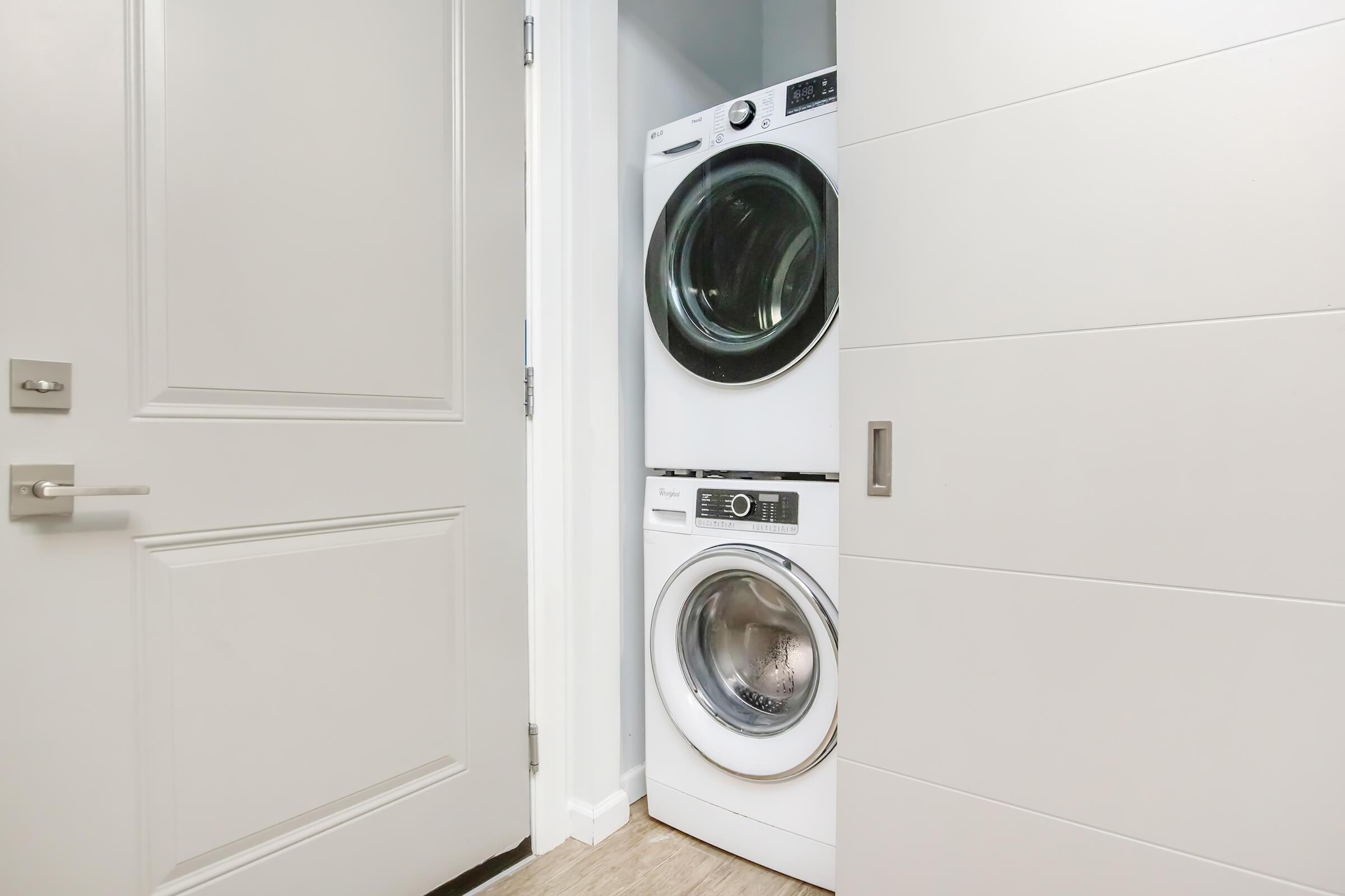 A modern laundry room featuring a stacked washer and dryer in white, positioned between a door and a wall with light-colored paneling. The space is bright and clean, with a minimalist design.