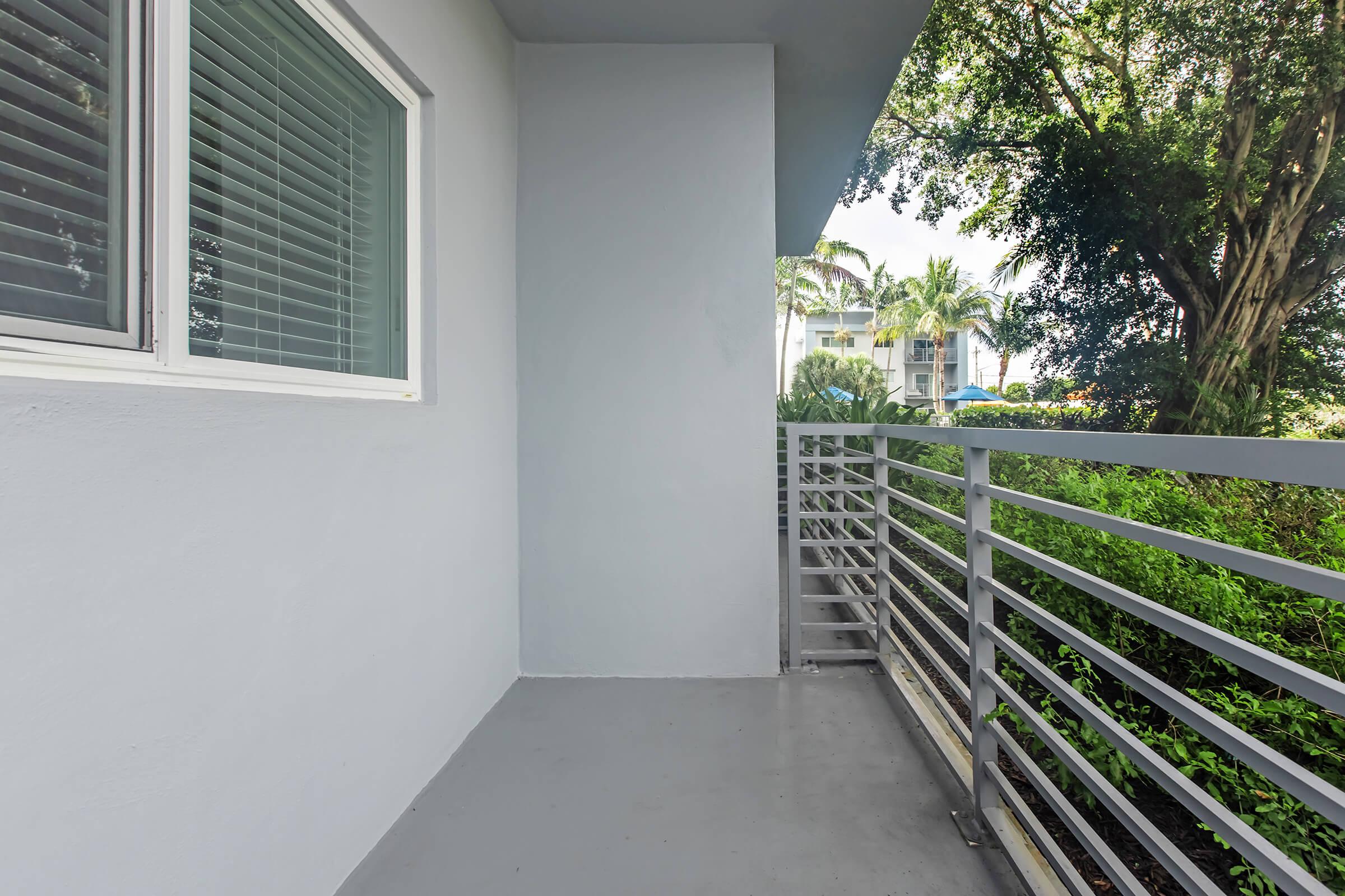 An empty balcony featuring a gray floor and railings, with a window on the left side. Lush greenery can be seen outside the railing, indicating a tropical environment. The space is well-lit, with sunlight filtering through the trees.