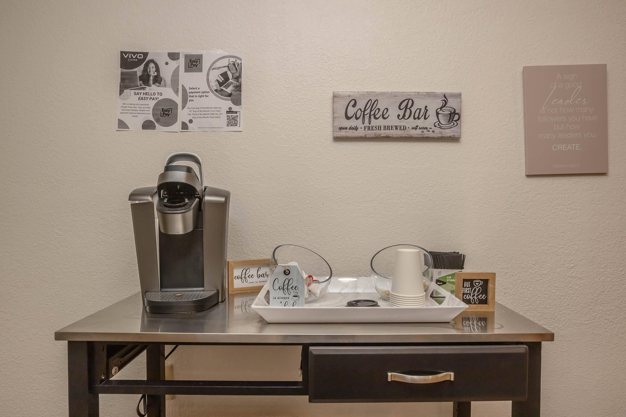 A coffee bar setup featuring a coffee machine, disposable cups, and a tray. Nearby, there are signboards and flyers mounted on the wall, advertising events and providing coffee-related information. The space is organized and inviting, perfect for guests to enjoy freshly brewed coffee.