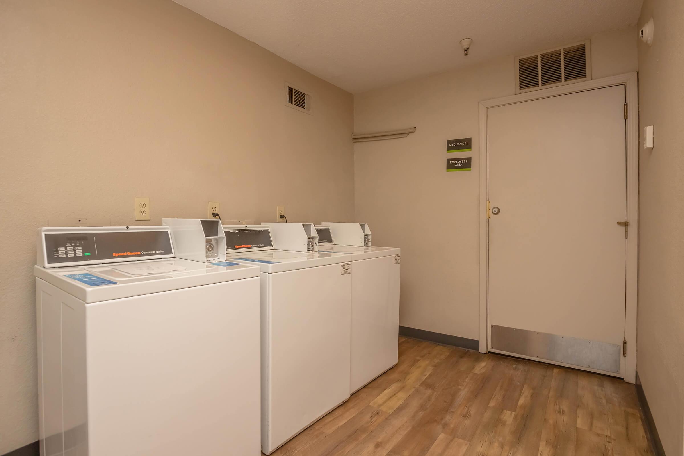 A small laundry room featuring three white washing machines lined up against a beige wall. There's a door at the end of the room, and a small shelf above the machines. The flooring is wooden, contributing to a simple, functional atmosphere.