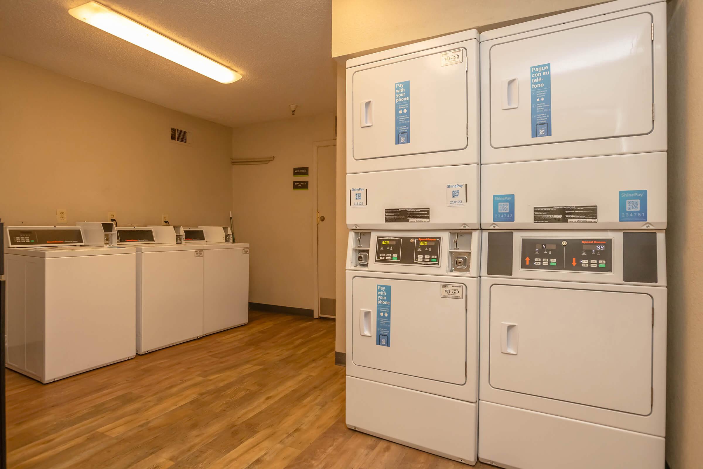 A laundry room featuring several white washing machines and dryers lined up against the wall. The machines are modern with digital controls, and the room has neutral-colored walls and wooden flooring. Soft lighting illuminates the space, creating a clean and functional environment for laundry tasks.