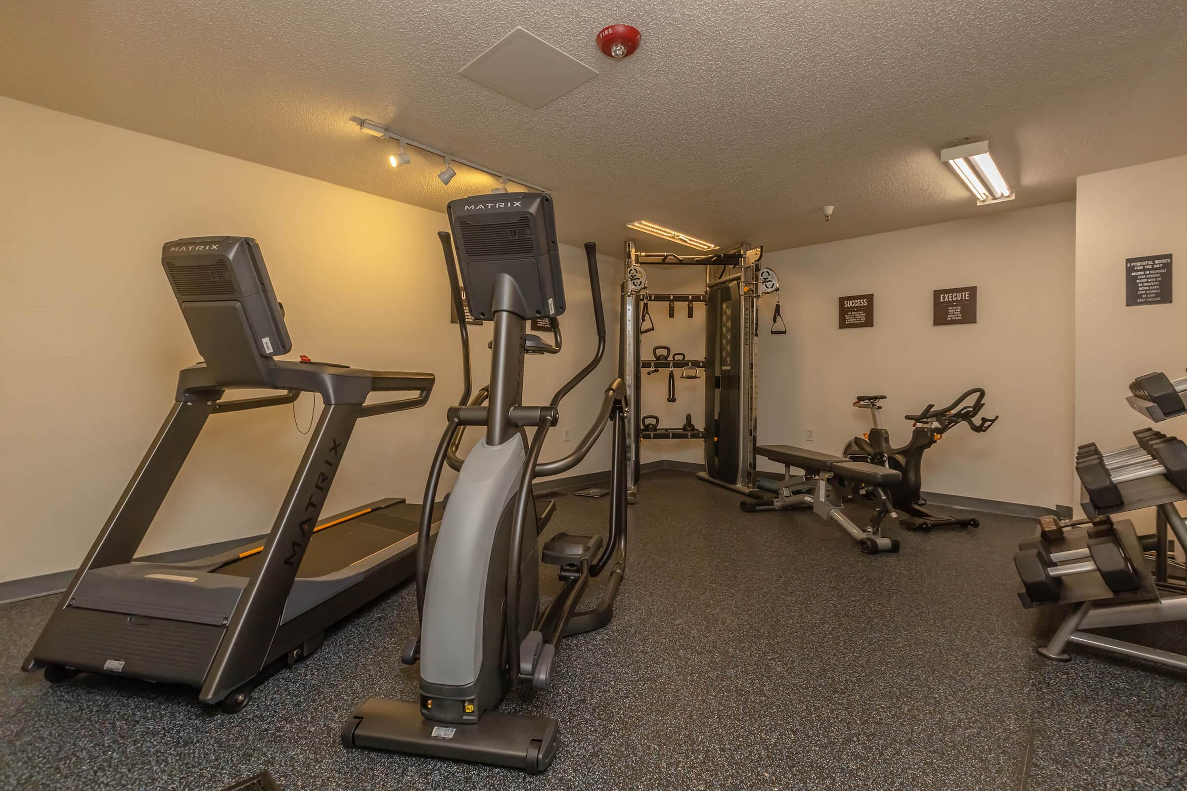 A well-equipped indoor gym featuring a treadmill, elliptical machine, stationary bike, multi-gym weight station, and free weights, with a clean and modern design, set against a neutral-colored wall and flooring.
