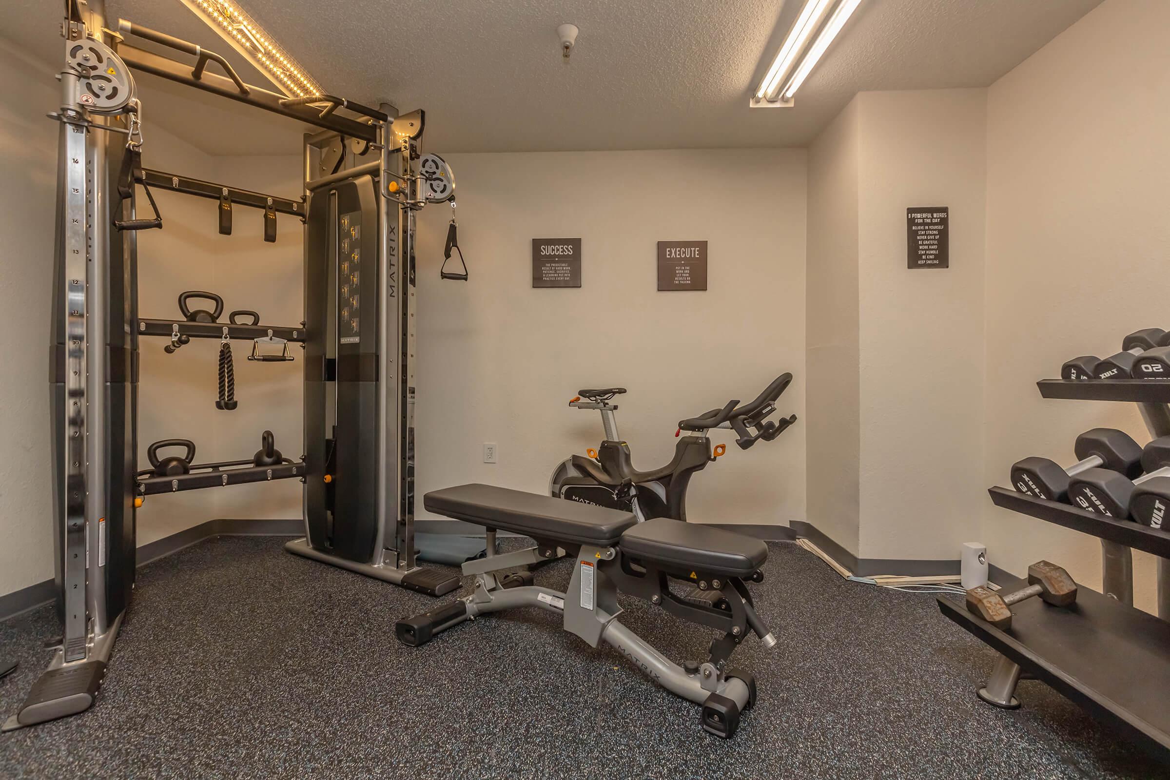 A fitness room featuring a multi-functional weight machine, a stationary bike, a weight bench, and a rack of dumbbells. The space has carpeted flooring and overhead lighting, with motivational wall signs visible in the background.