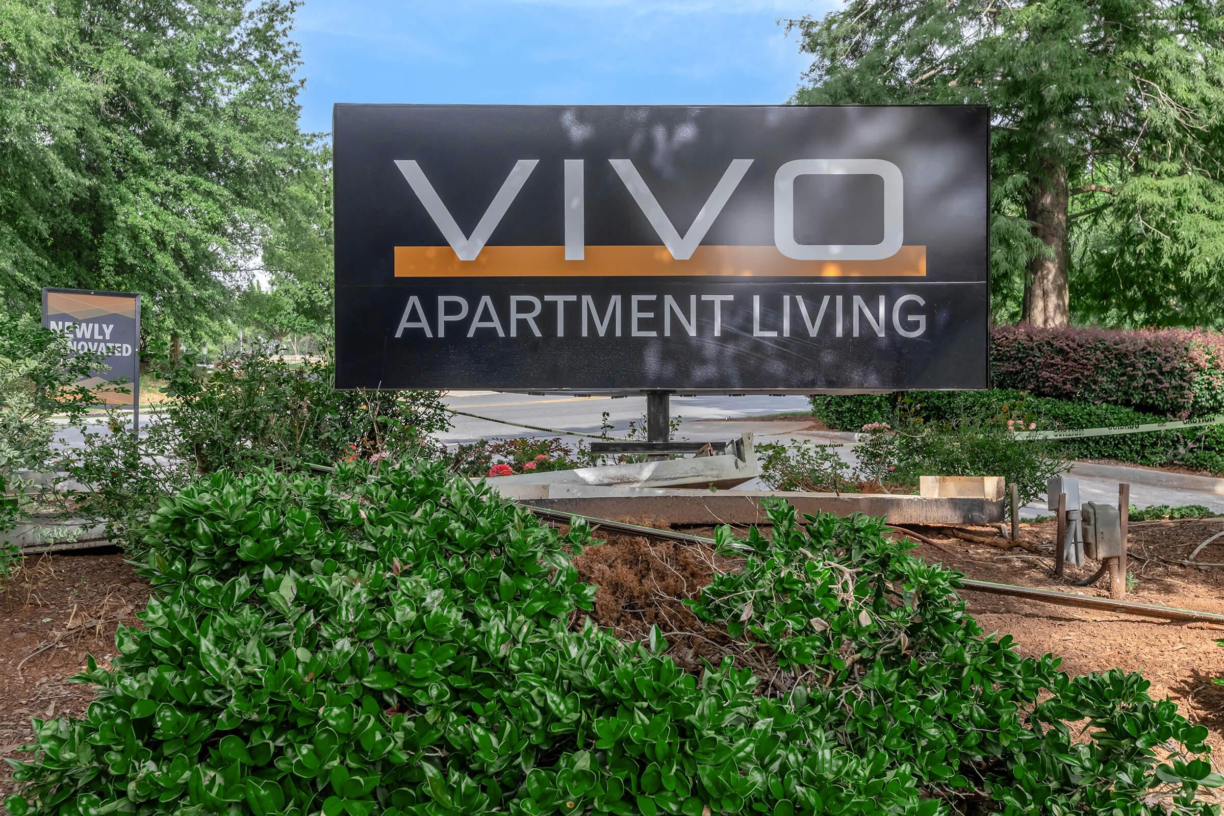 Sign for "VIVO Apartment Living" prominently displayed, surrounded by greenery. The sign indicates a modern apartment complex, with a note that the apartments are "Newly Renovated." The background features trees and landscaping typical of a residential area.