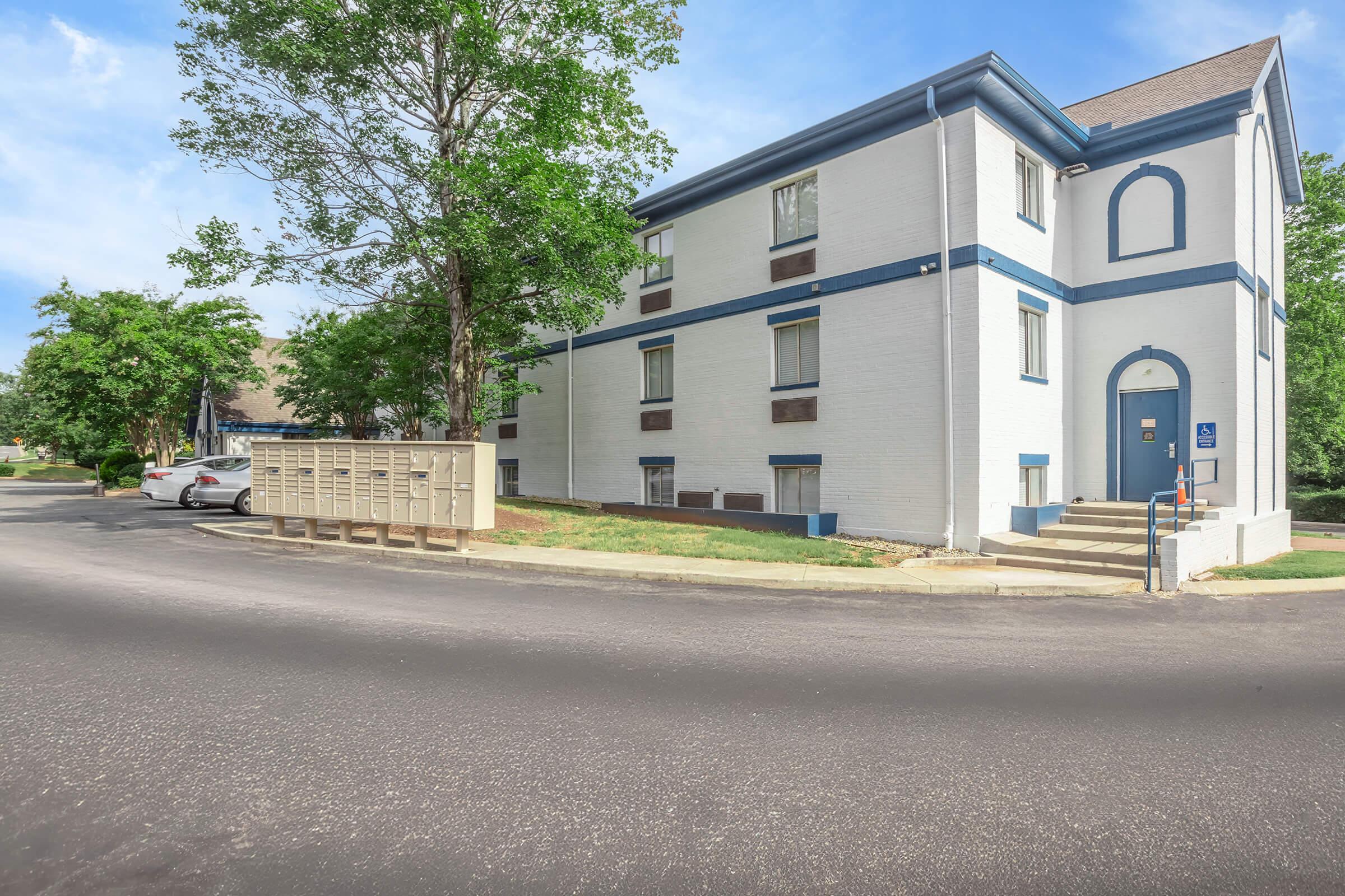 A three-story white apartment building with blue accents surrounded by greenery. There is a parking area in front, and a set of mailboxes is visible near the entrance. The scene is set on a sunny day with a clear blue sky.