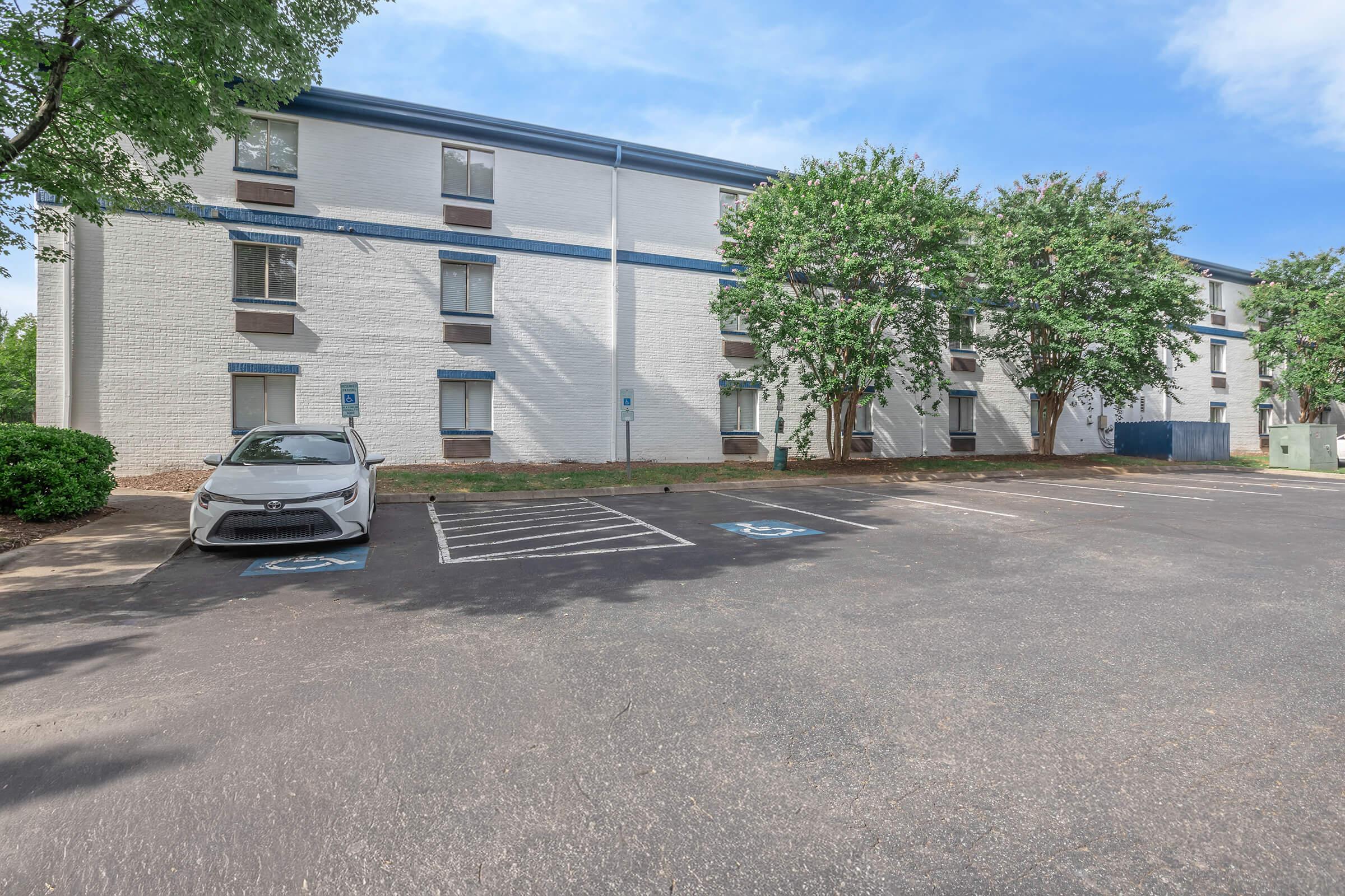 A view of a multi-story building with a light-colored exterior, surrounded by trees. In the foreground, there is a parking lot with a white car parked in a designated spot. The area appears well-maintained with a clear blue sky above.