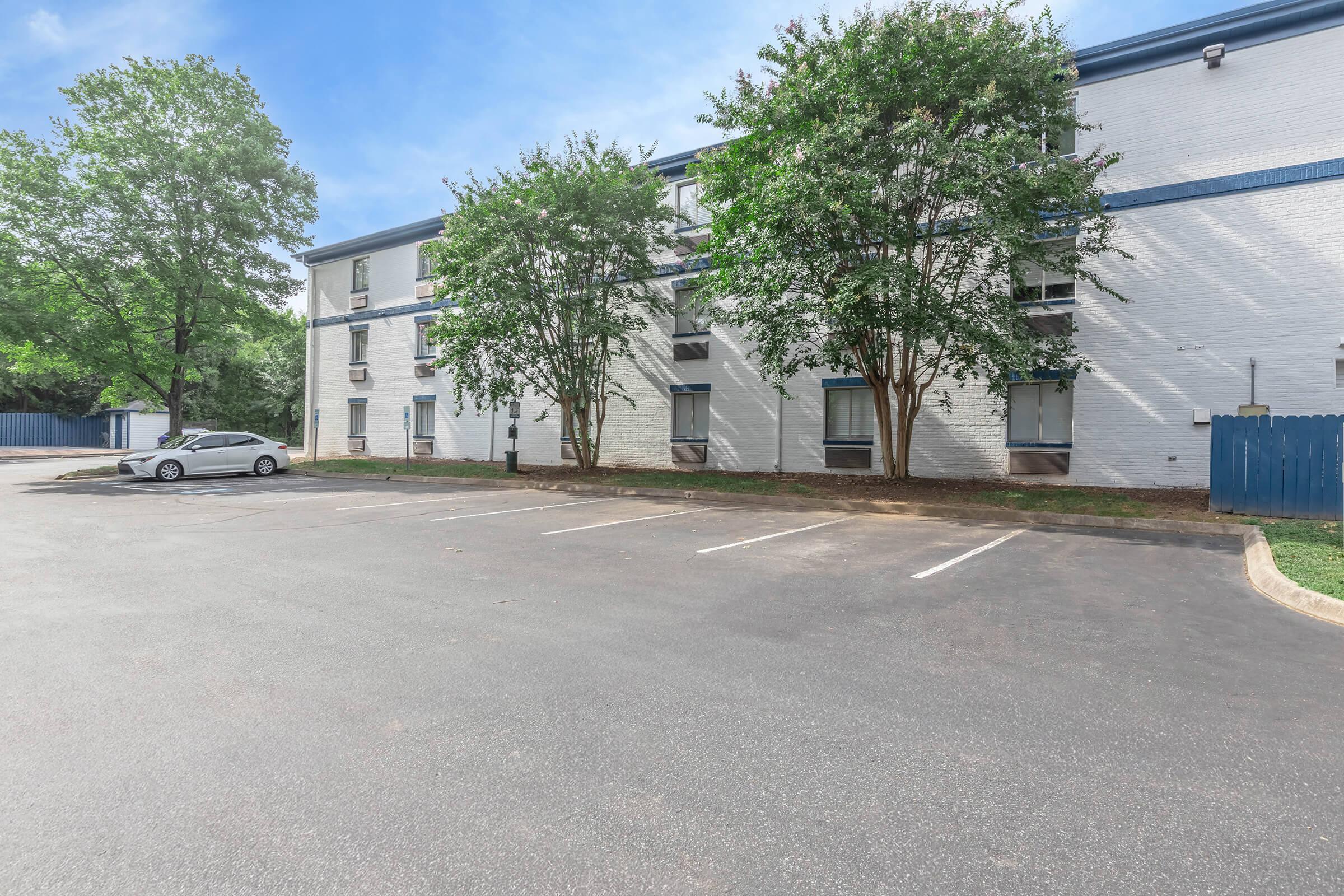 View of a three-story motel-style building with a white exterior, surrounded by green trees. An empty parking lot is visible in front, with a single car parked. The sky is clear and blue, and a wooden fence is partially obscured by foliage in the background.