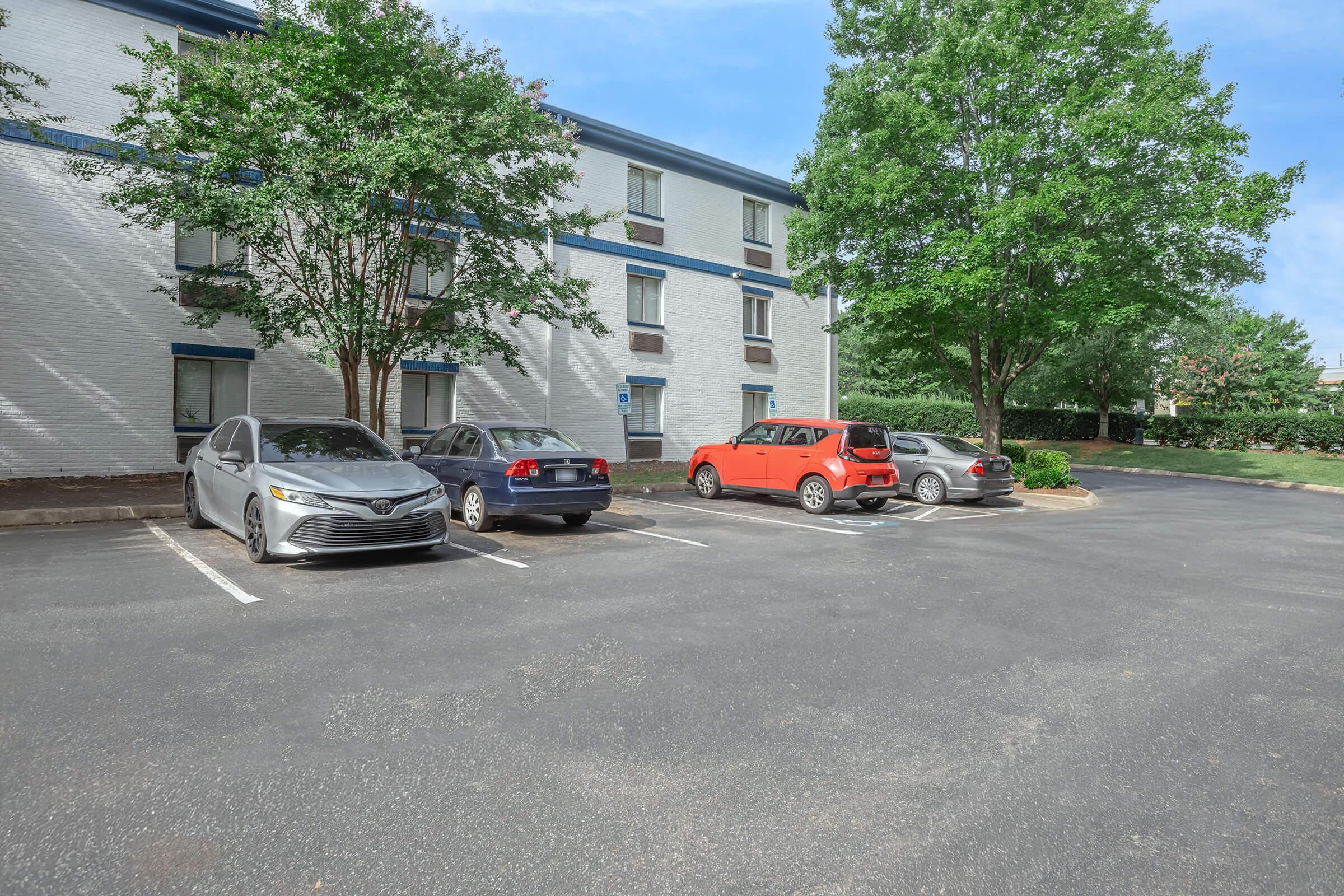 A parking lot featuring four cars parked in front of a multi-story building surrounded by green trees and grass. The building has white walls with blue accents.