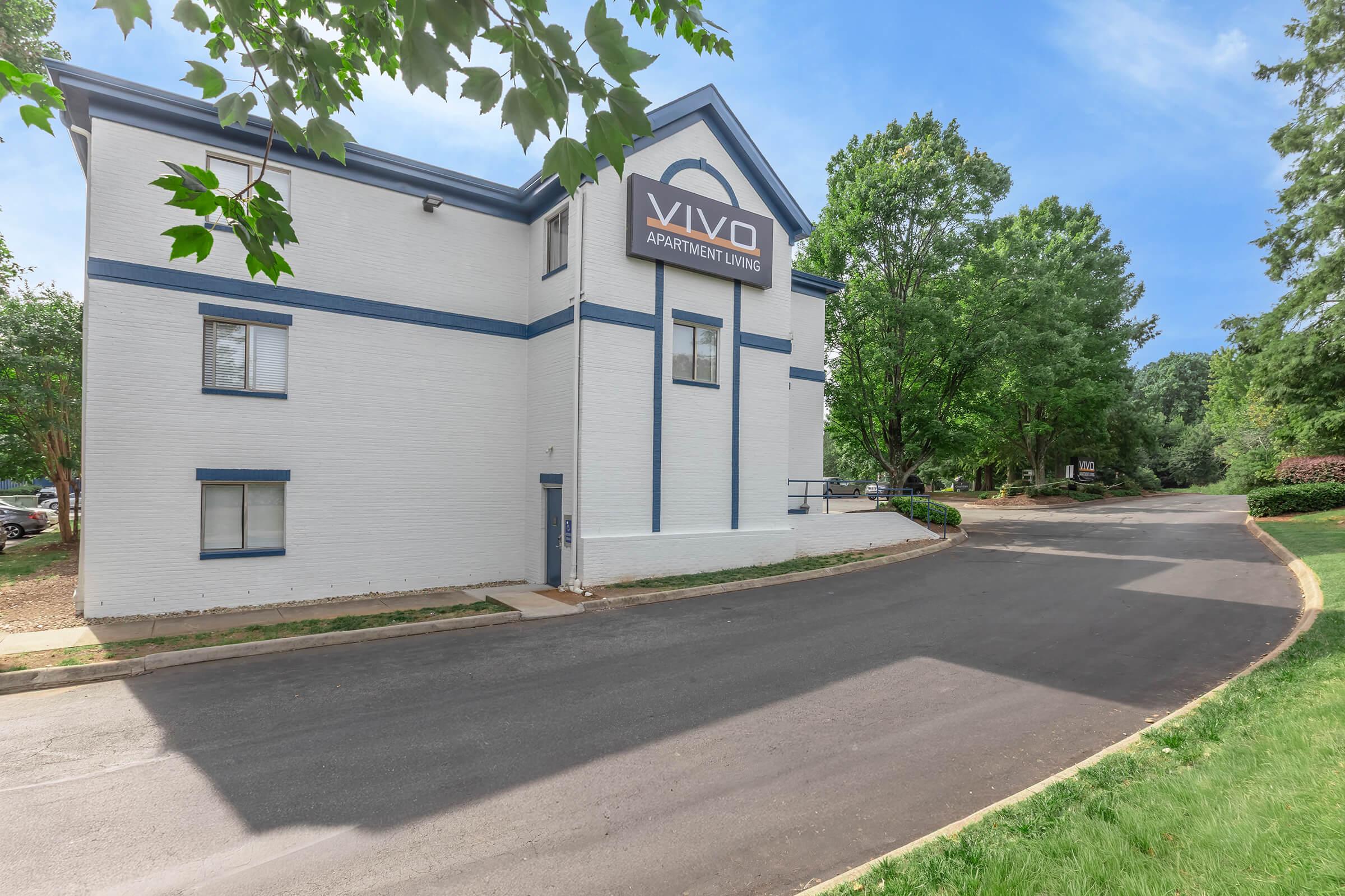 VIVO Apartment Living building exterior featuring a two-story structure with white brick and blue accents, surrounded by greenery. A paved road leads up to the entrance, with windows visible on both floors. The signage displays "VIVO APARTMENT LIVING" prominently.