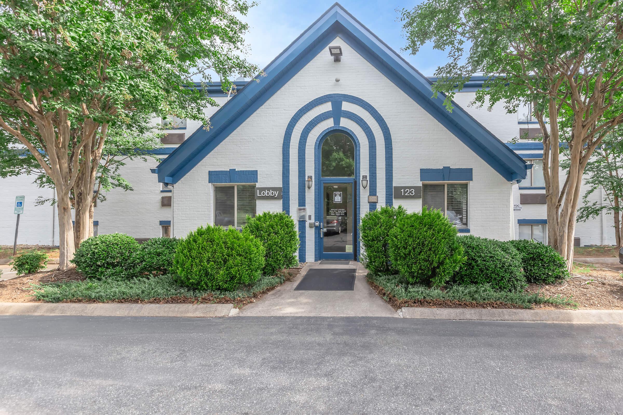 A well-maintained building featuring a blue and white exterior design. The entrance shows a lobby sign and is flanked by neatly trimmed bushes. Two trees are positioned on either side of the walkway leading to the door. The overall appearance is inviting and well-kept.