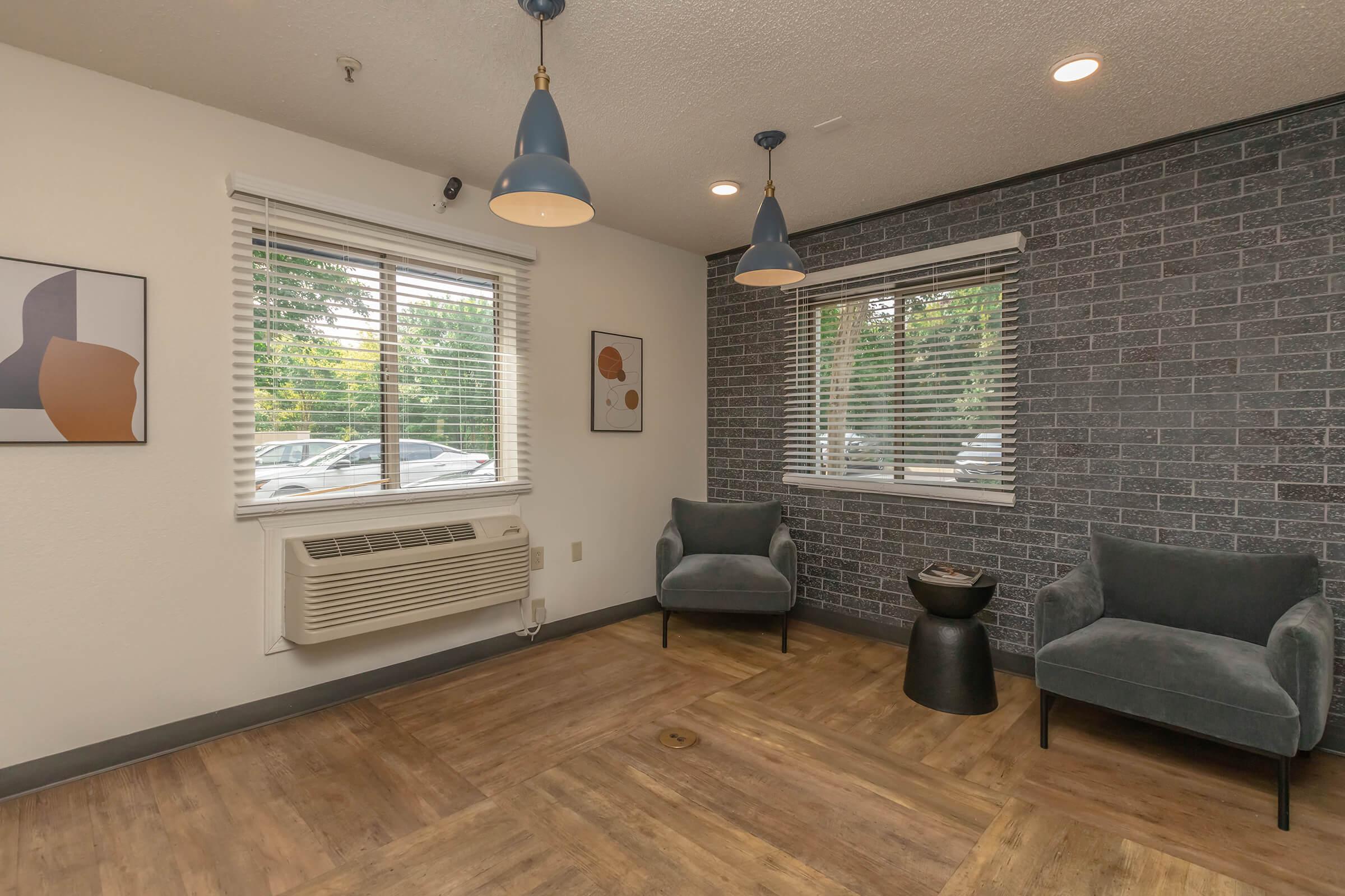 A cozy interior space featuring two gray armchairs, a small black side table, and a window with blinds. Wall decor includes a framed abstract art piece. The room has laminate flooring and a gray brick accent wall, with stylish pendant lighting hanging from the ceiling.