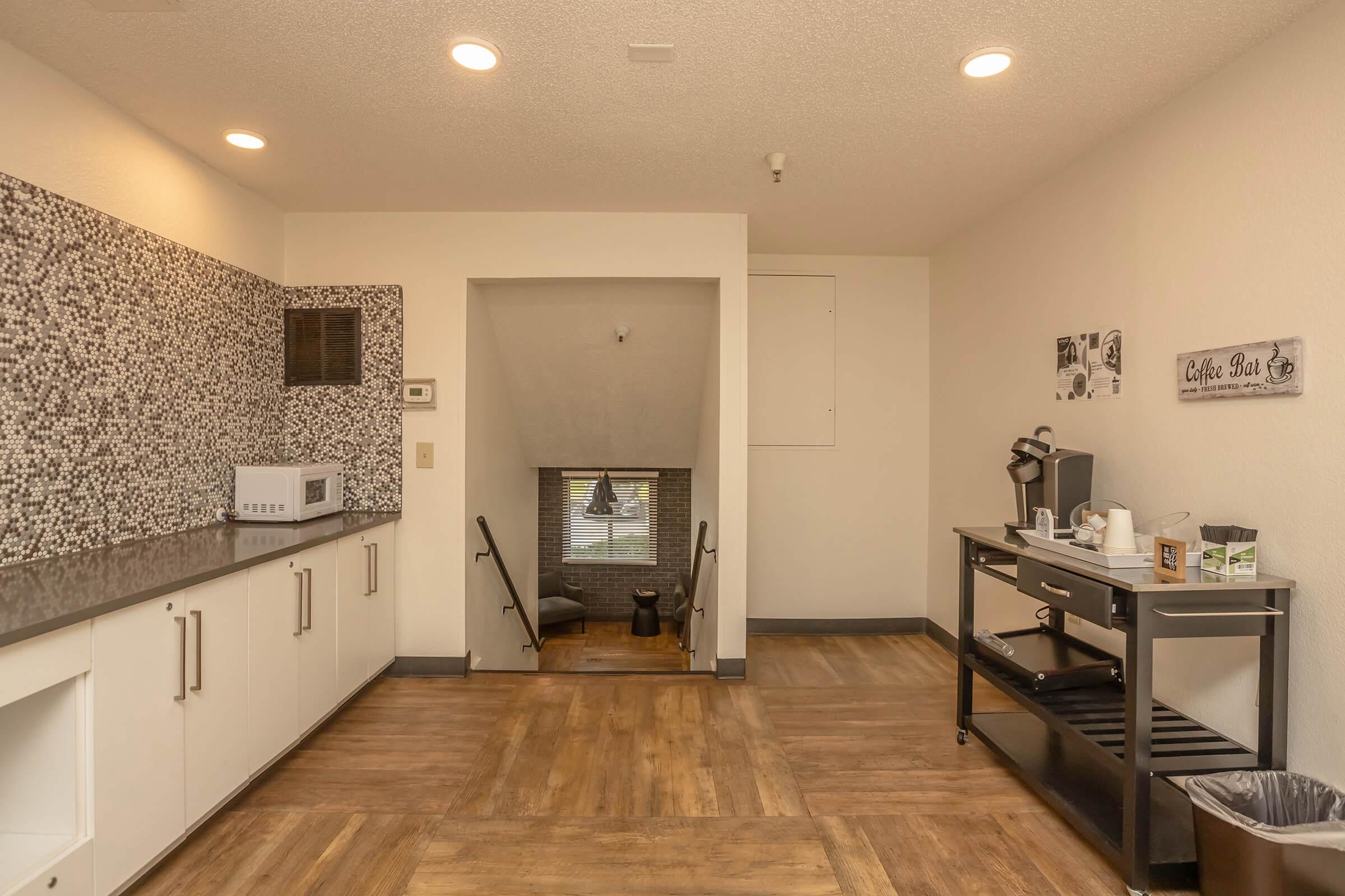 A modern kitchenette with a dark countertop, microwave, and coffee maker. The space features a mosaic wall and warm lighting. In the background, a staircase leads down to a lower level, and there's a black cart on wheels for additional storage. The flooring is a light wood finish.