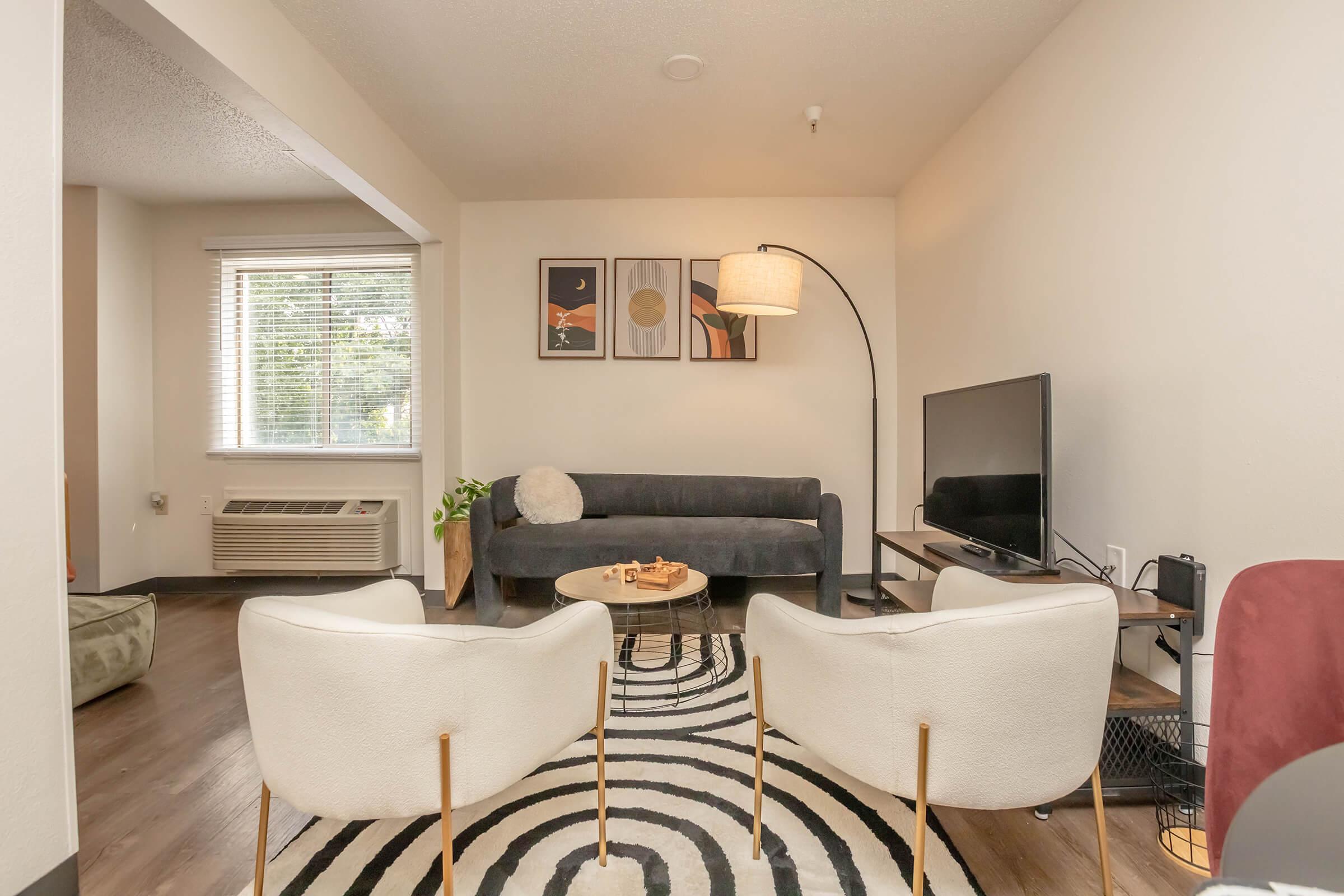 A cozy living room featuring a dark gray sofa, two white armchairs, and a round coffee table with decorative items. A large flat-screen TV sits on a wooden stand, and a stylish floor lamp is nearby. The room is decorated with abstract wall art and has a patterned rug on the floor. Natural light filters through a window.