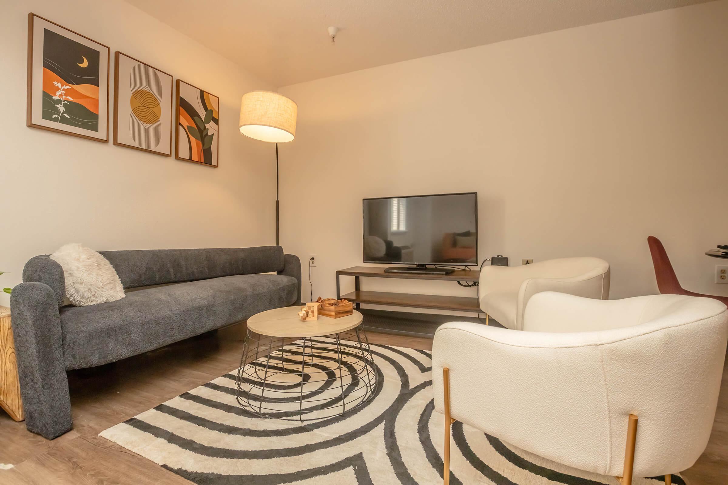A cozy living room featuring a gray sofa with a textured throw, two white accent chairs, a circular coffee table with a small game, and a flat-screen TV on a wooden stand. The space is decorated with three framed artwork pieces on the wall and a floor lamp providing warm lighting, all set on a stylish area rug.