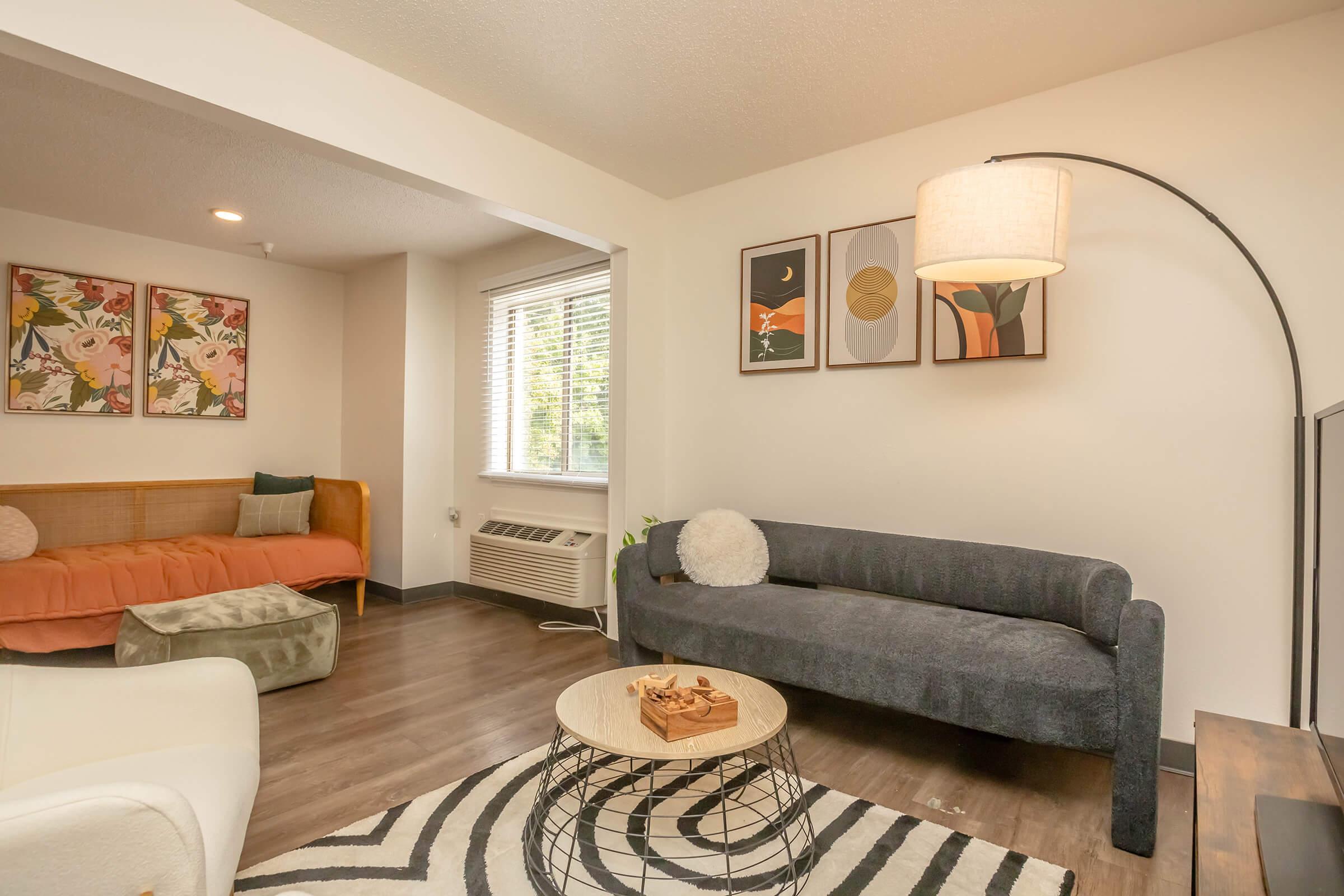 A modern living room featuring a gray sofa, a round coffee table with a decorative item, a light-colored armchair, and an orange daybed. The walls are adorned with colorful artwork, and there is a large window allowing natural light to illuminate the space. A stylish floor lamp stands in the corner.