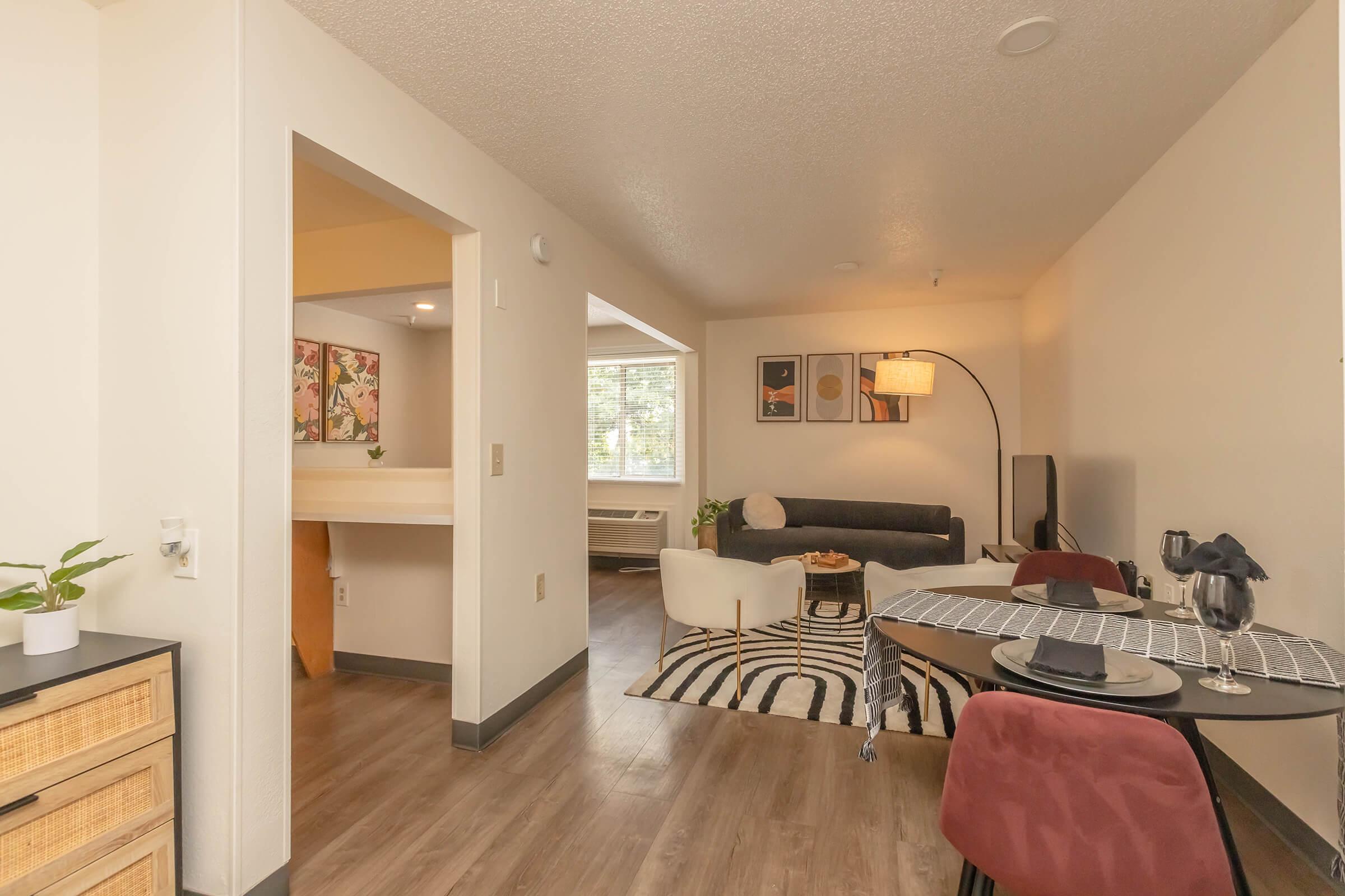 A modern living space featuring a cozy sitting area with a dark couch, a round coffee table, and a stylish floor lamp. There is a dining table with chairs, a patterned rug on the floor, and decorative artwork on the walls. Natural light enters through a window, creating a welcoming ambiance.