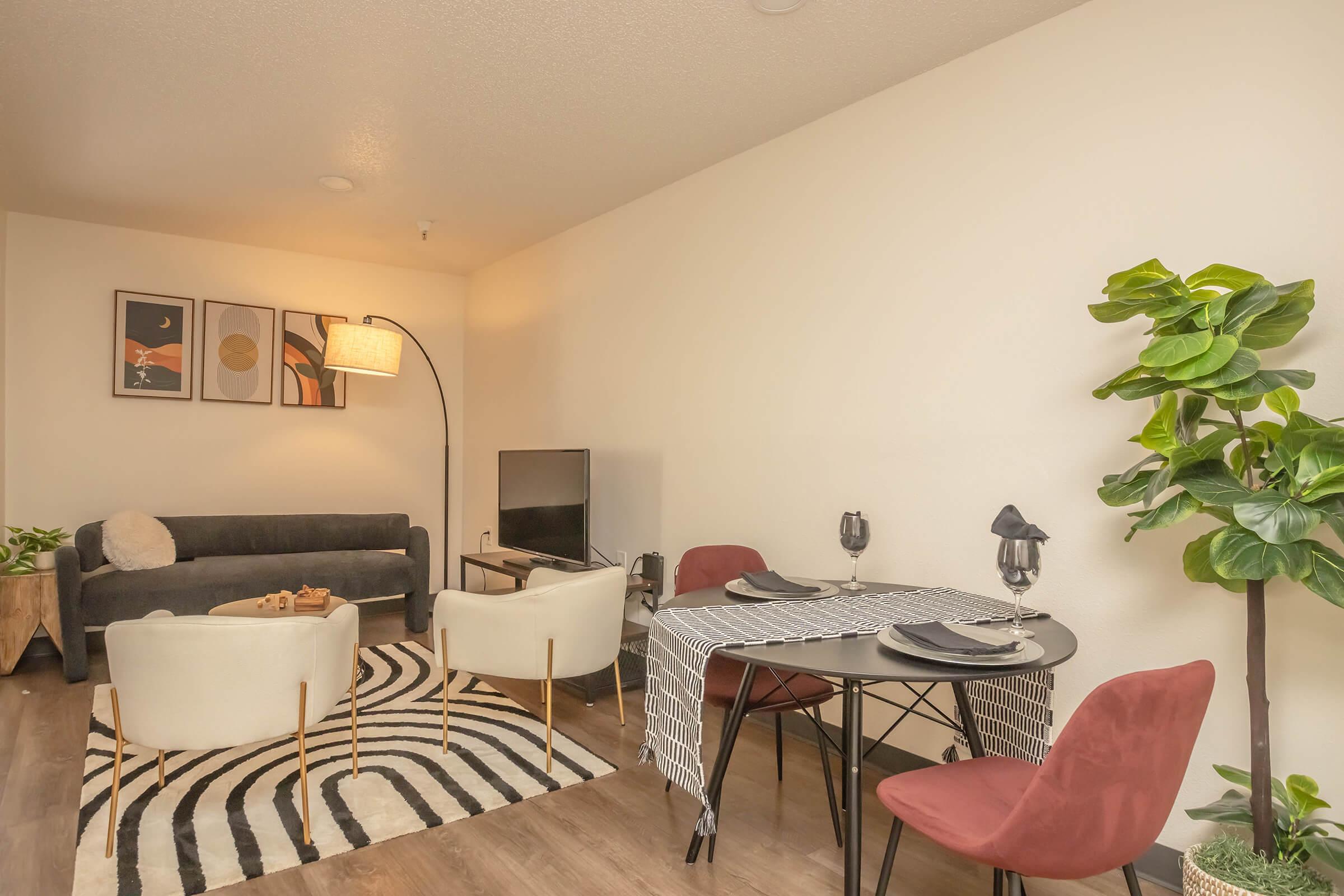 A modern living space featuring a dark sofa, two stylish chairs, a dining table set for two, and a TV. The room has a large floor lamp, decorative artwork on the walls, and a potted plant, with a black and white striped rug underfoot. The decor is warm and inviting.
