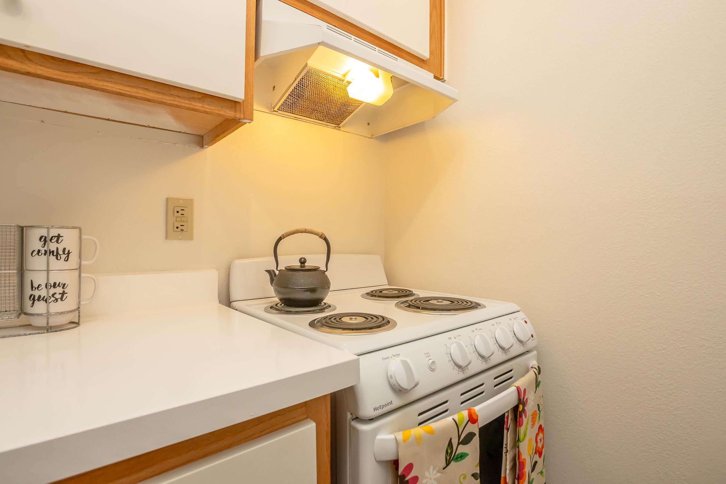 A small kitchen with a white stove, a kettle on the burner, and a lit range hood. There is a white countertop and wooden cabinets above. A decorative towel hangs from the stove. A coffee mug with text is visible on the countertop. The wall is painted in a light color, creating a cozy atmosphere.