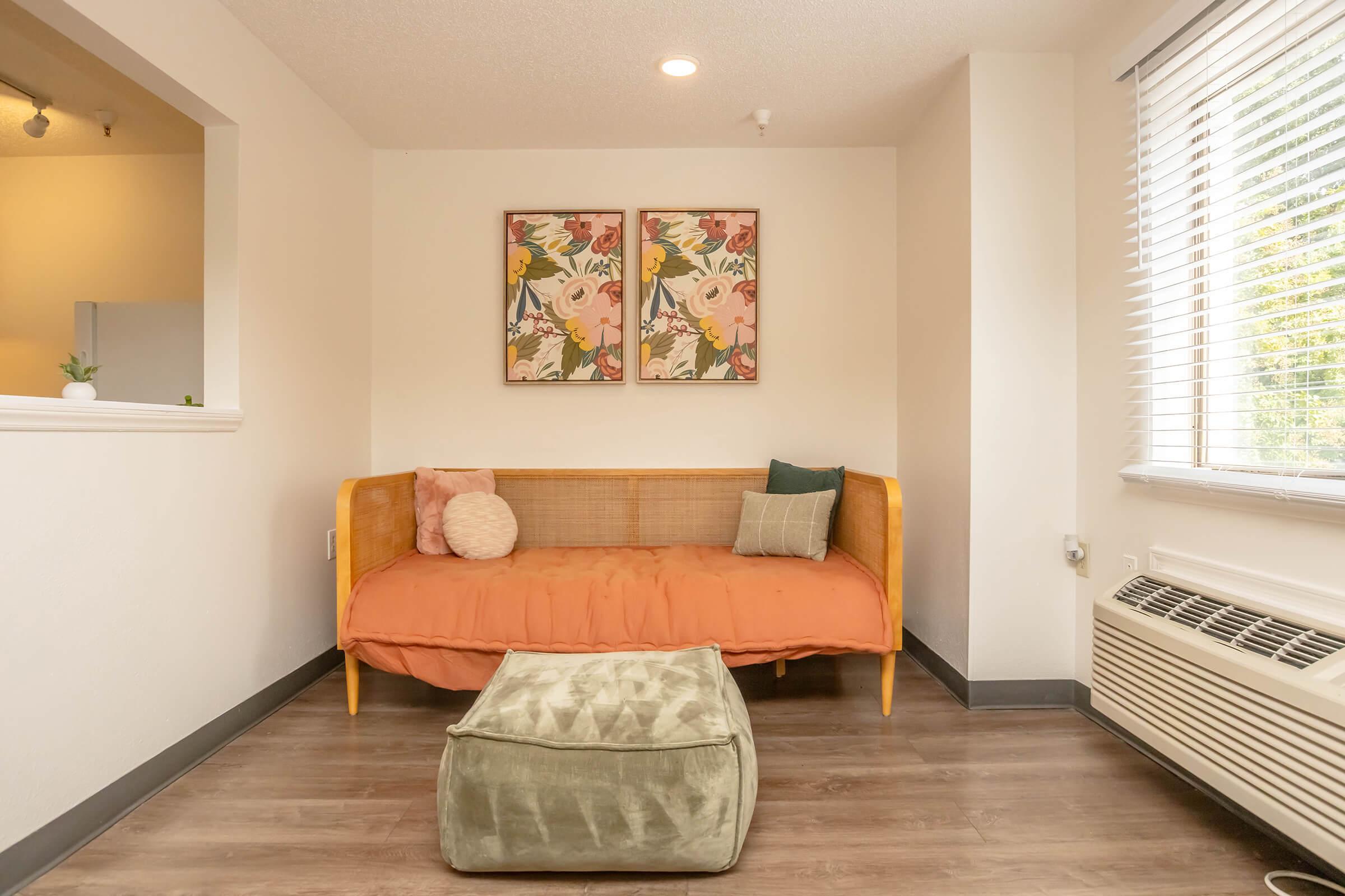 A cozy living space featuring a small orange couch with decorative cushions, a green ottoman, and two framed floral art pieces on the wall. Natural light streams in through a window with blinds, and the floor has a wooden appearance. A subtle, modern aesthetic is evident throughout the room.