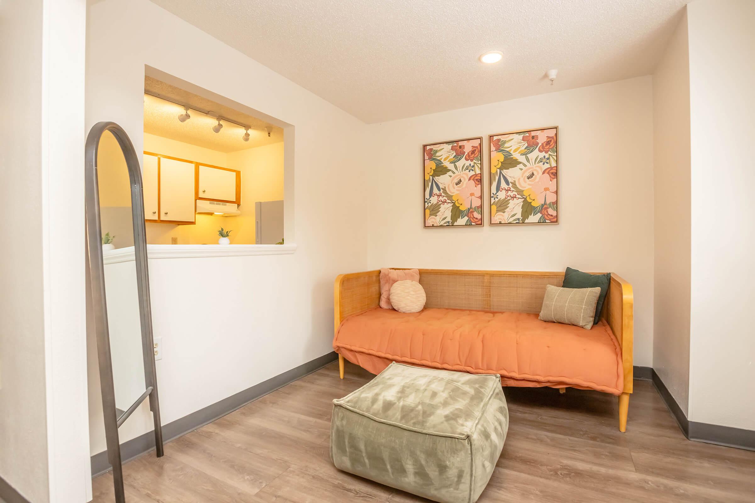 A cozy living space featuring a wooden daybed with orange bedding, accompanied by soft pillows and a green ottoman. Two floral-themed framed artworks adorn the wall above the daybed. A glimpse of a kitchen area with light cabinets can be seen in the background. The flooring is a warm wood finish, enhancing the inviting atmosphere.