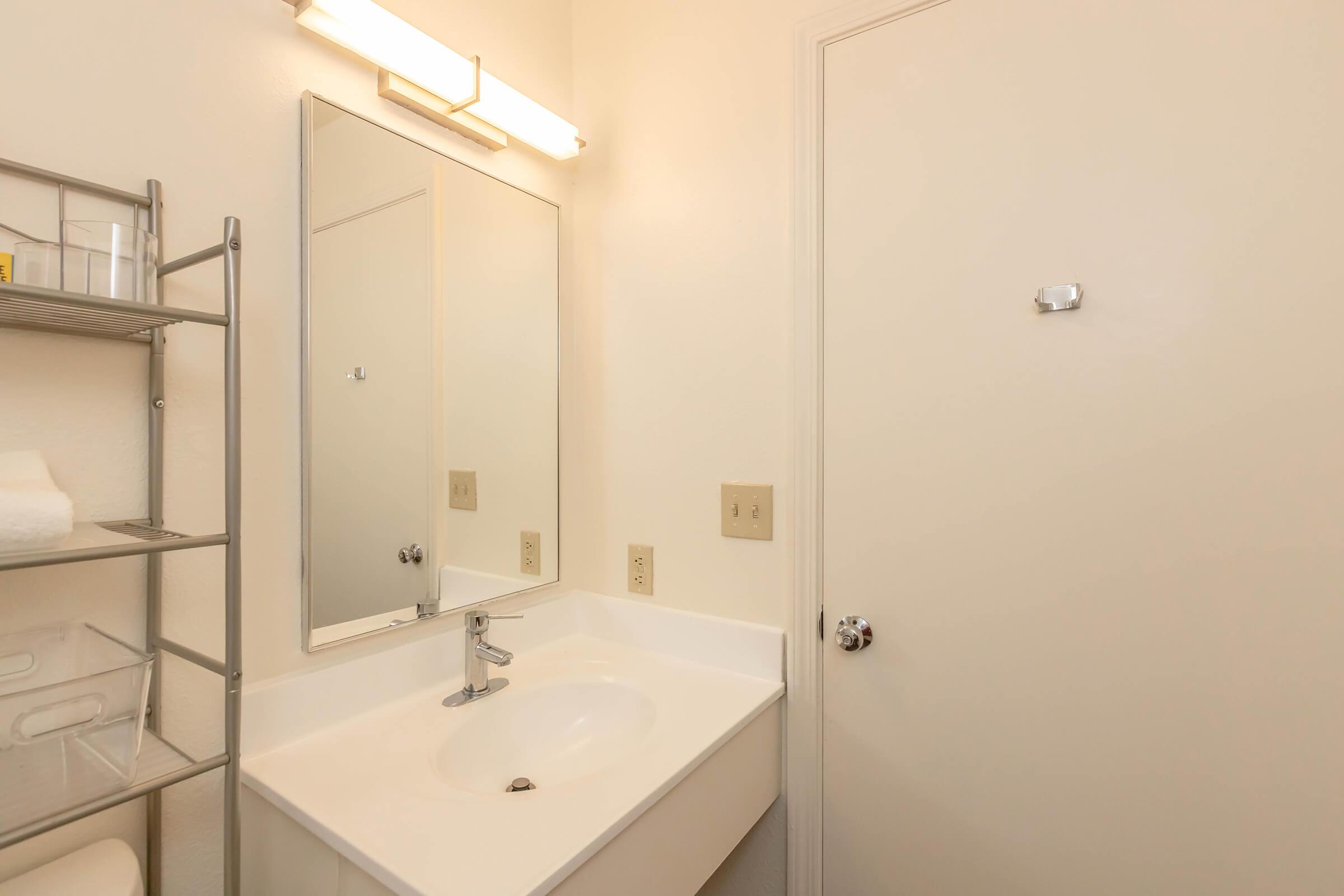 A small bathroom featuring a white sink, a wall-mounted mirror, and a door. The walls are light-colored, and there is a towel rack with white towels nearby. A light fixture is above the mirror, providing illumination. Simple and clean design with minimal decorations.