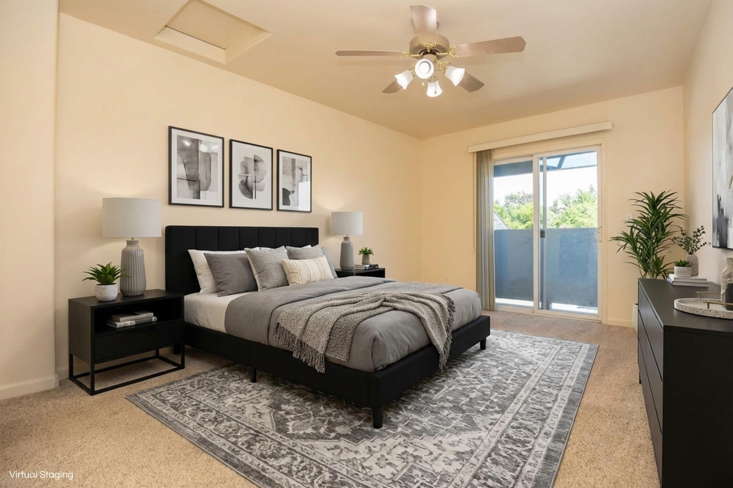 A modern bedroom with a large bed dressed in gray and white linens, flanked by two bedside tables with lamps. Features include a dark dresser, a patterned area rug, and three framed pictures on the wall. A sliding glass door leads to a balcony, and plants add a touch of greenery to the space.