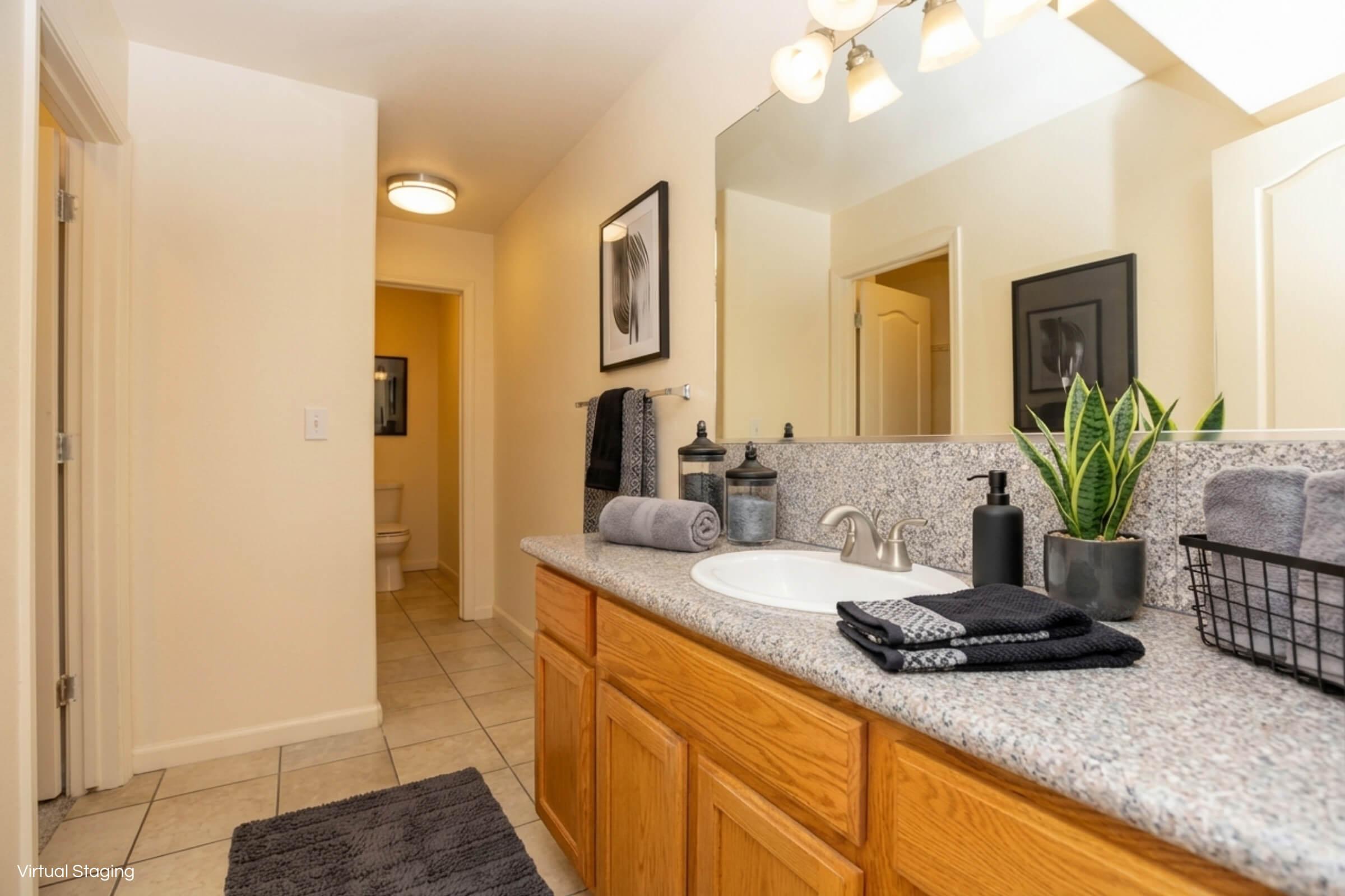 A well-lit bathroom featuring a double vanity with a granite countertop, dark towels, and decorative items. There is a potted plant and a mirror above the sink. A door leads to a separate toilet area in the background. The overall decor is modern and clean, with neutral tones and careful organization.