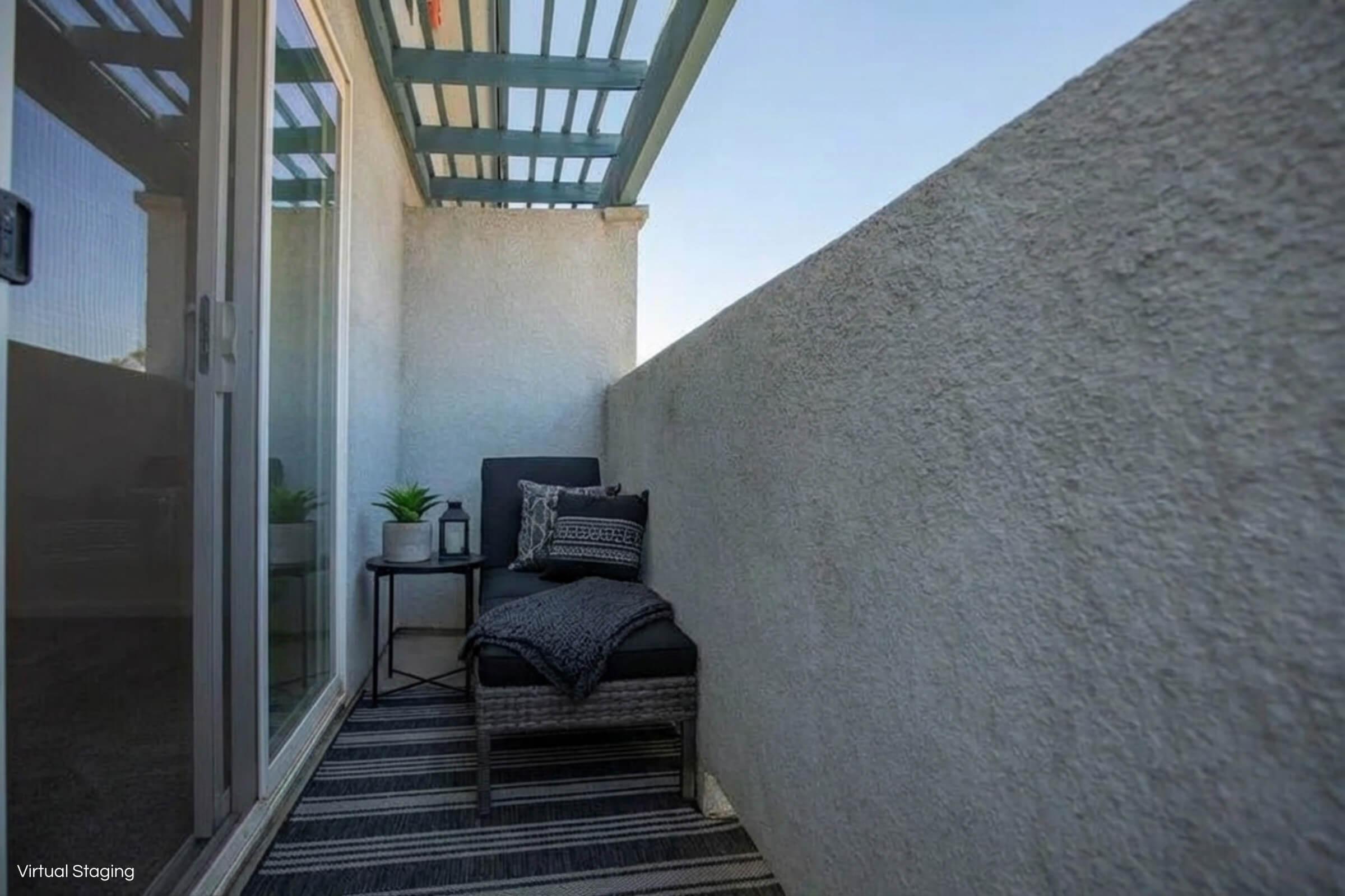 A small balcony with a modern design features a comfortable chair with a cozy throw blanket and a side table. The space is adorned with a potted plant, and the walls are painted in neutral tones. A woven rug adds warmth, and sunlight filters through a slatted overhead structure, creating a serene atmosphere.