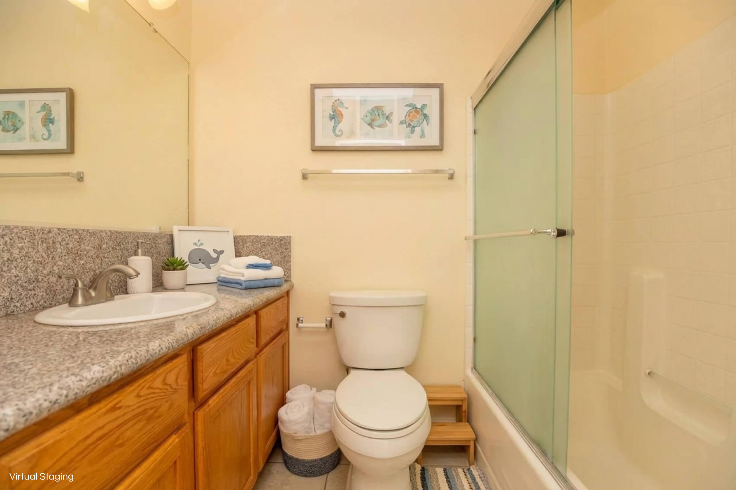 A clean and modern bathroom featuring a white toilet, a granite countertop with a sink, and a glass shower enclosure. The space is decorated with framed seahorse artwork and includes a small plant on the counter, towels stacked nearby, and a wooden shelf for additional storage.