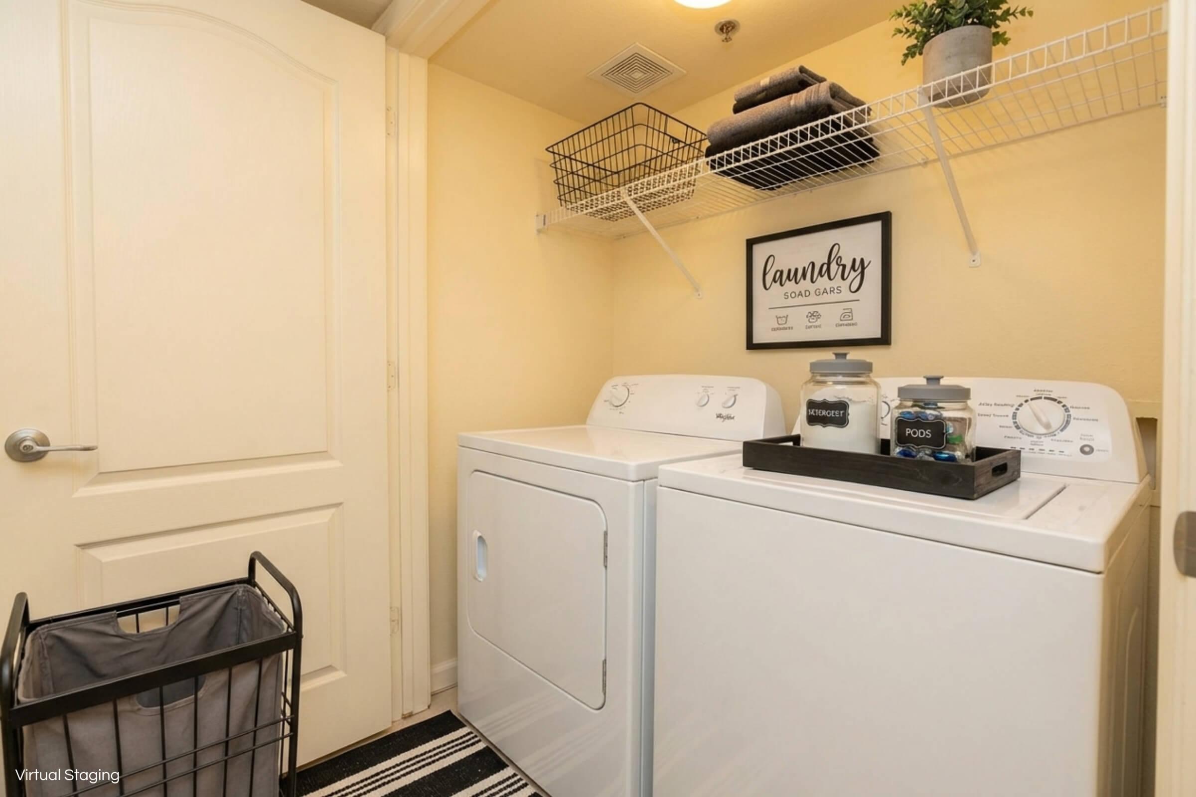 A small laundry room featuring a white washing machine and dryer side by side. Above them, a shelf holds neatly stacked towels and a decorative sign that reads "Laundry." Two glass jars labeled "Detergent" and "Pods" are placed on top of the dryer. A gray laundry basket sits nearby on a striped rug.