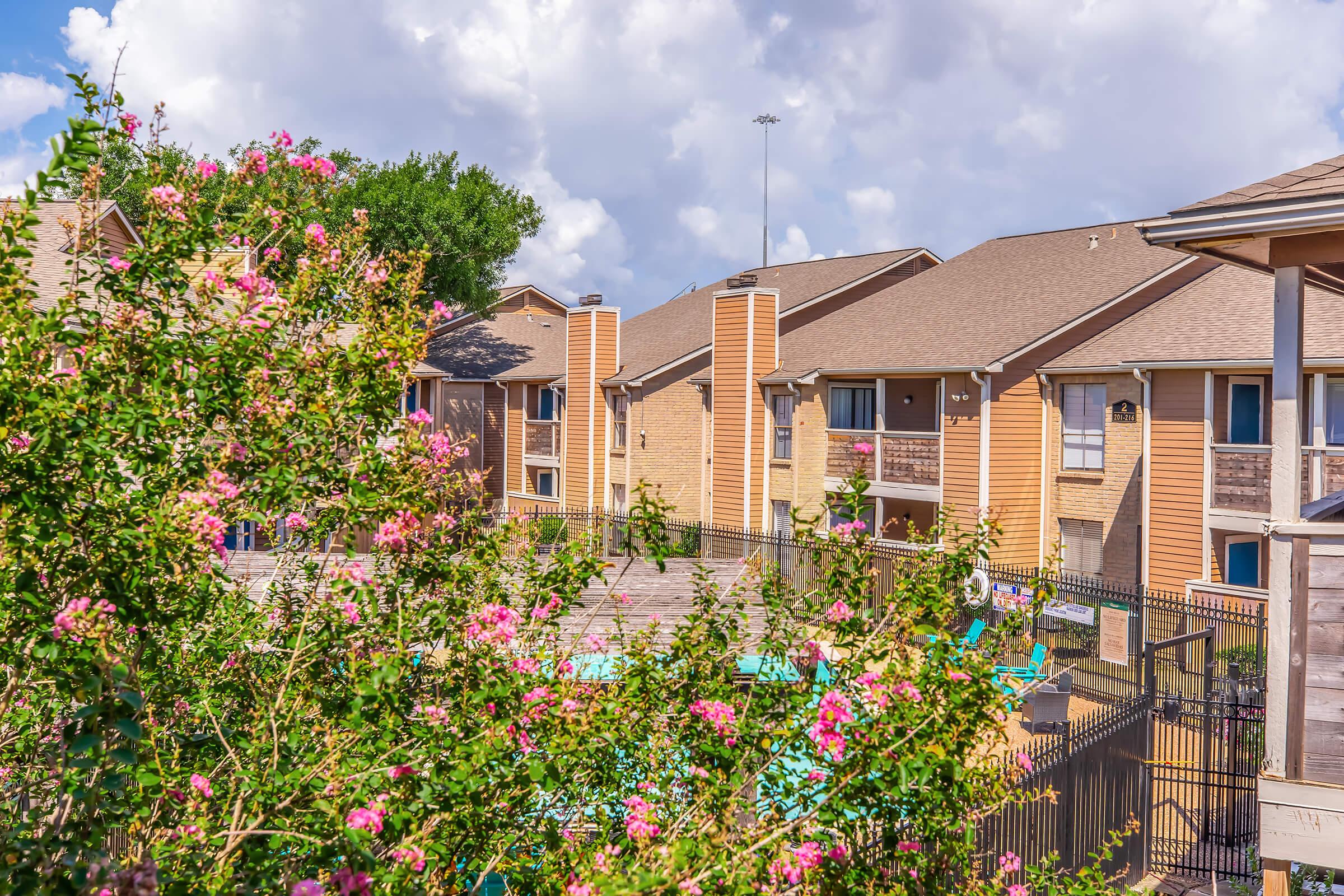 A view of an apartment complex featuring tan buildings with balconies, surrounded by vibrant pink flowers and green shrubs. A swimming pool is visible in the courtyard area, set against a backdrop of a cloudy blue sky.
