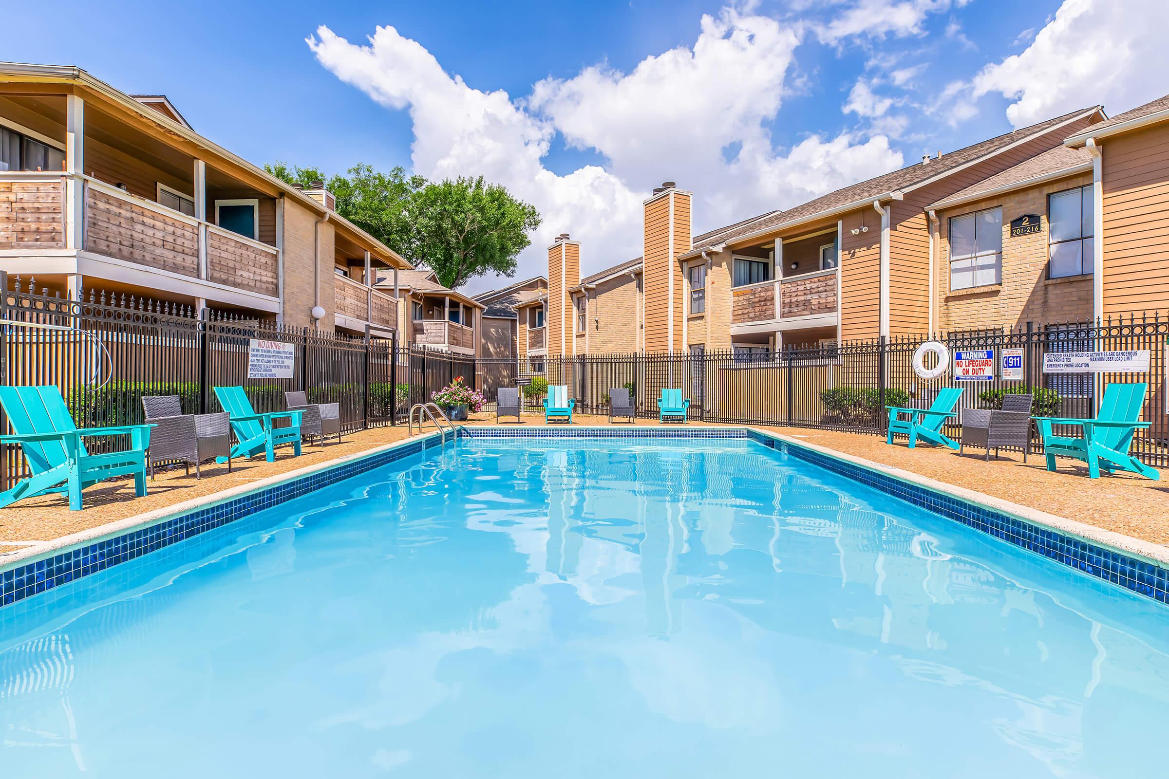 A bright and clear swimming pool surrounded by lounge chairs, with a backdrop of two-story apartment buildings and lush greenery. The sky is mostly sunny with a few fluffy clouds, creating a welcoming atmosphere for relaxation and leisure by the poolside.