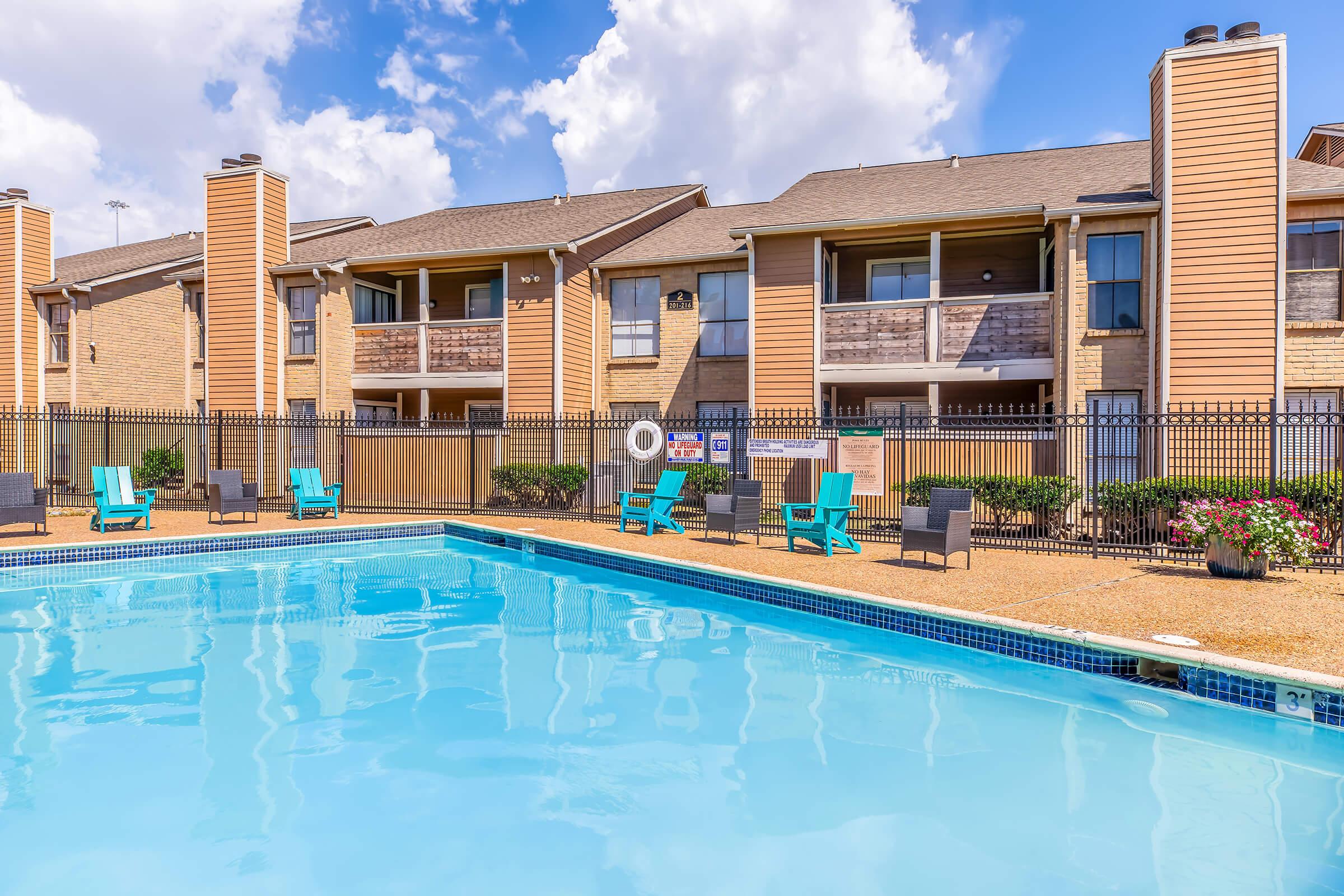 A sparkling blue swimming pool surrounded by lounge chairs and tables, with a view of two-story apartment buildings in the background. The scene is bright and sunny, featuring fluffy white clouds in a blue sky, creating a relaxing outdoor atmosphere.