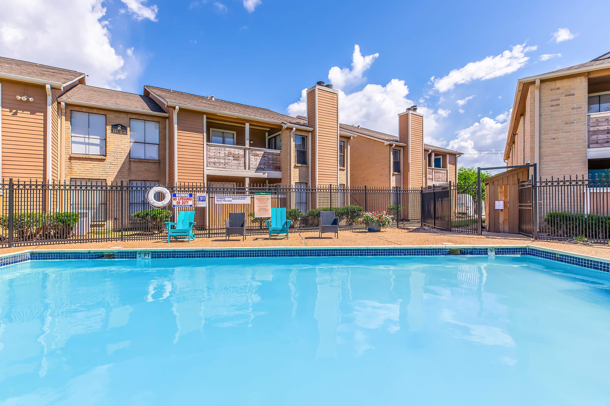 A clear swimming pool surrounded by a fenced area, with lounge chairs and a sunny blue sky dotted with clouds. In the background, there are two multi-unit residential buildings with balconies. The landscape is well-maintained with some greenery.