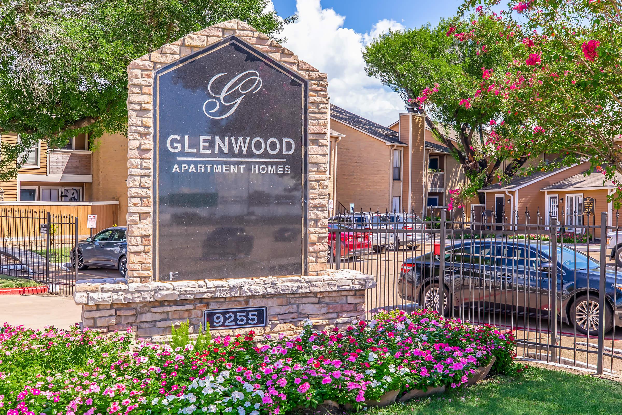 A stone sign for Glenwood Apartment Homes, featuring the logo and name "Glenwood" prominently displayed. The sign is surrounded by colorful flower beds and greenery, set against a blue sky with fluffy clouds. Nearby, several cars are parked in front of the apartment buildings.