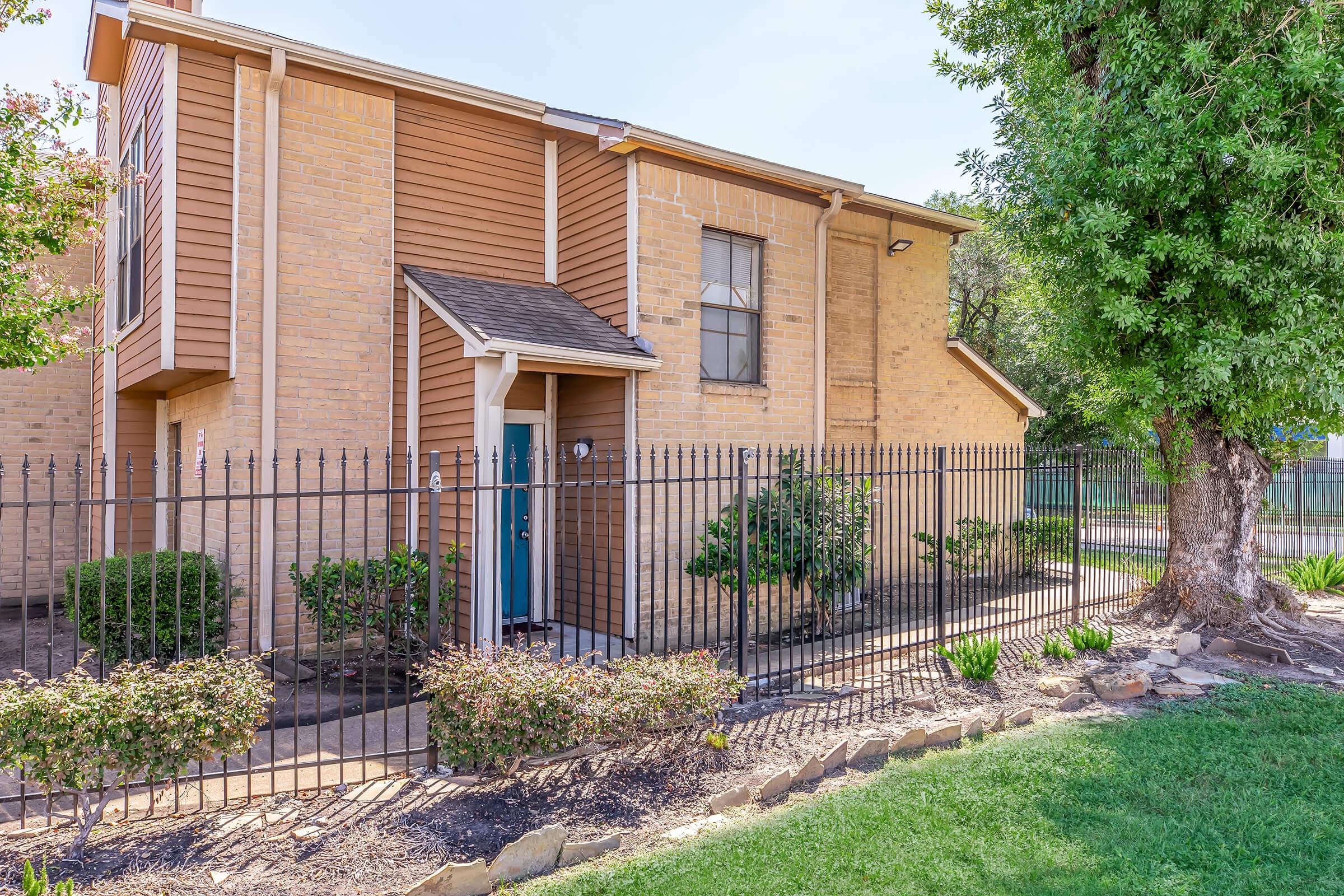 A two-story building with orange-brown brick siding and a blue door, surrounded by a black metal fence. There are small bushes and green plants in the garden area, with a large tree partially visible on the right. The setting appears to be sunny and well-maintained, indicating a residential area.