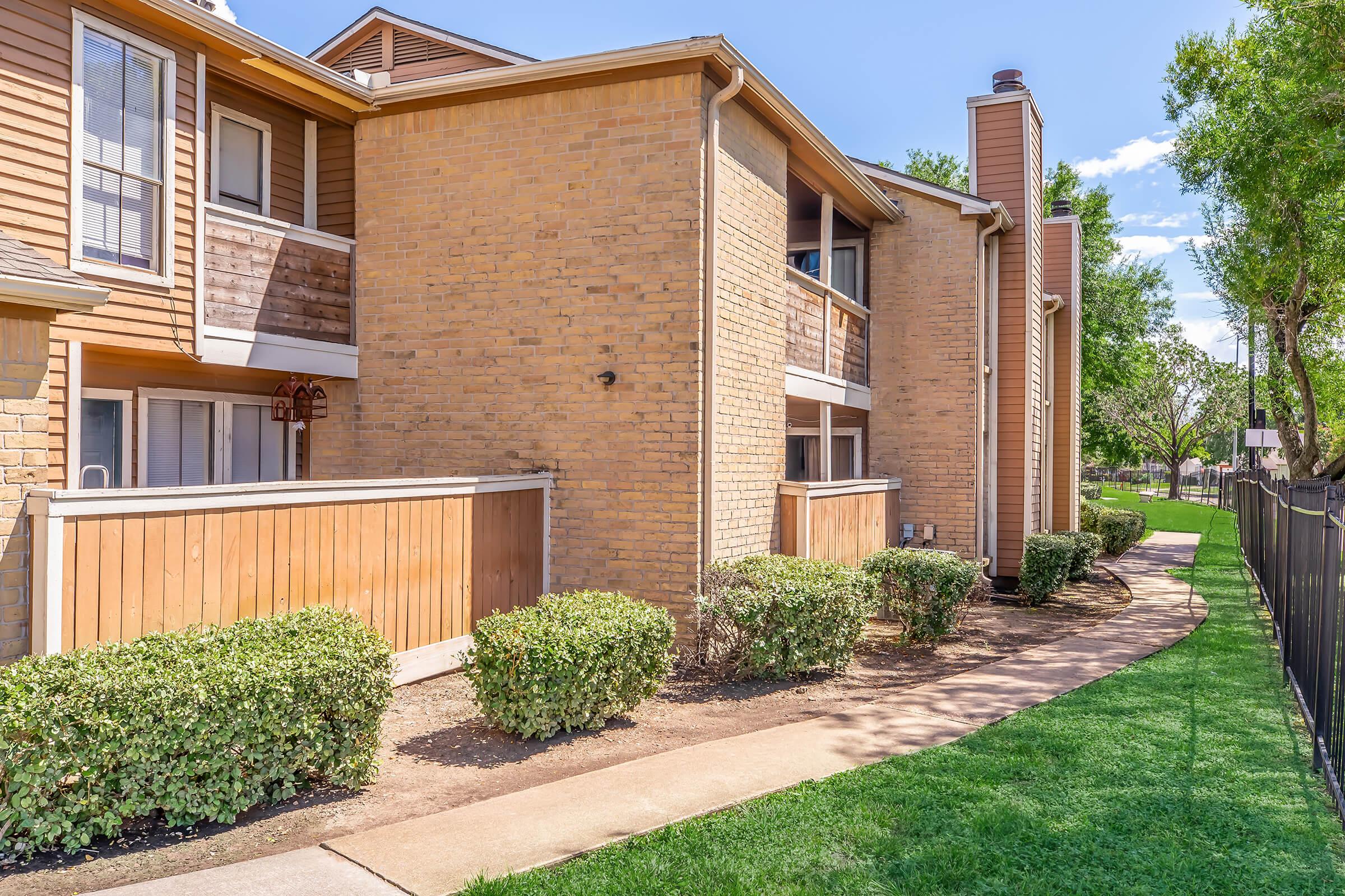 A corner view of a residential building with brick exterior and multiple balconies. Pathway lined with small shrubs leads to the entrance, with a well-maintained lawn and a fence visible in the background. Blue sky and trees can be seen, indicating a sunny day.