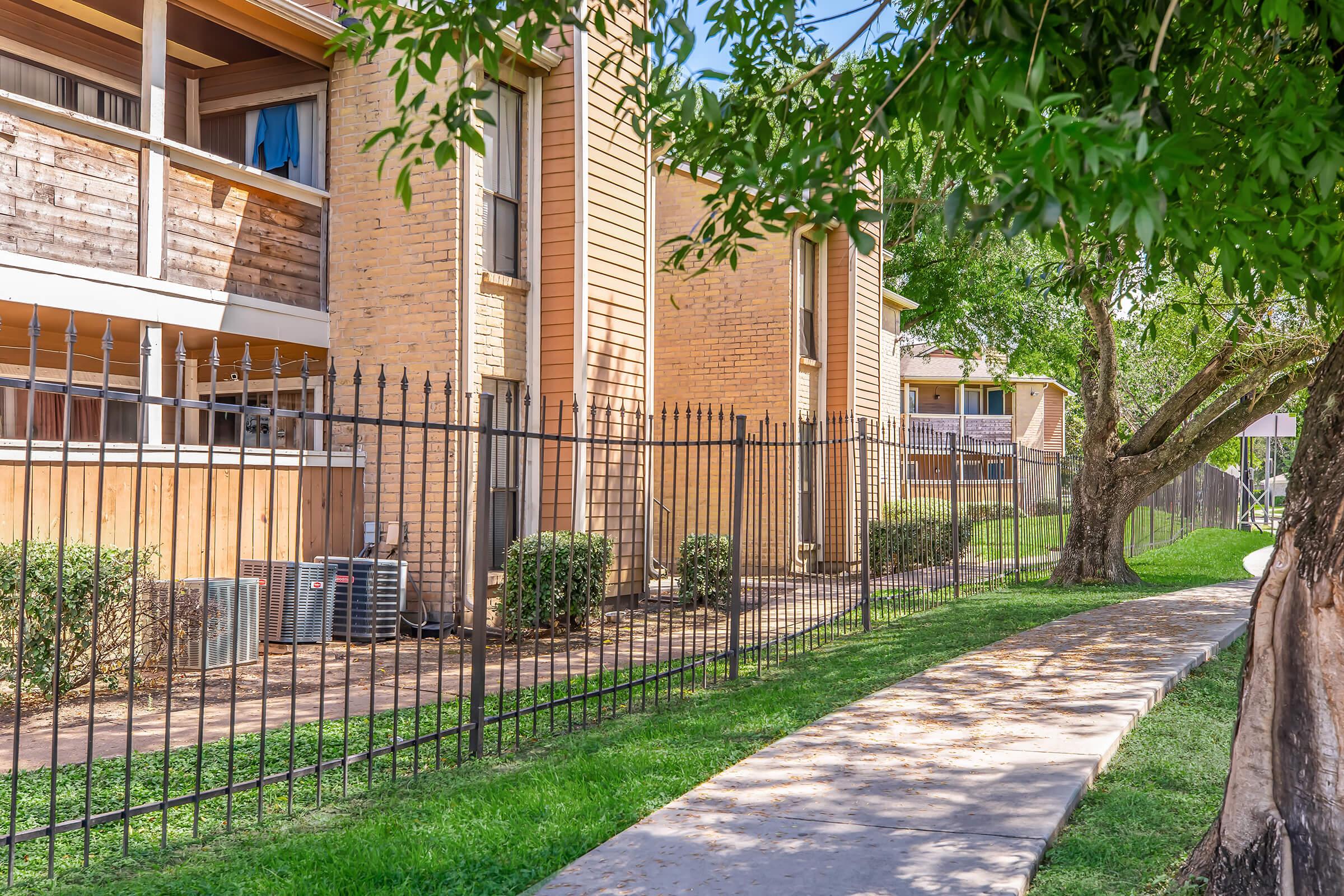 A view of an apartment complex featuring beige brick buildings with balconies and a black iron fence. In the foreground, a paved walkway runs alongside a green lawn with shrubs. The scene is shaded by a tree, indicating a pleasant outdoor environment.