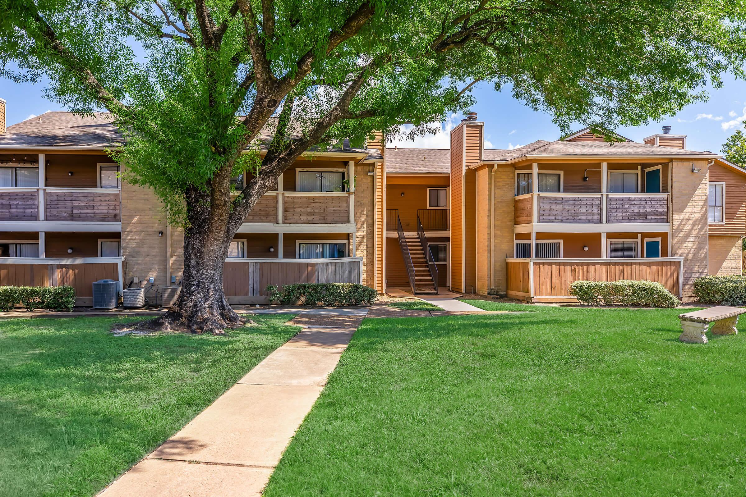A well-maintained apartment complex featuring two adjacent buildings with balconies. They are surrounded by lush green lawns and a large tree, creating a pleasant outdoor space. A concrete pathway leads through the yard, connecting the buildings. Clear blue skies add to the inviting atmosphere.