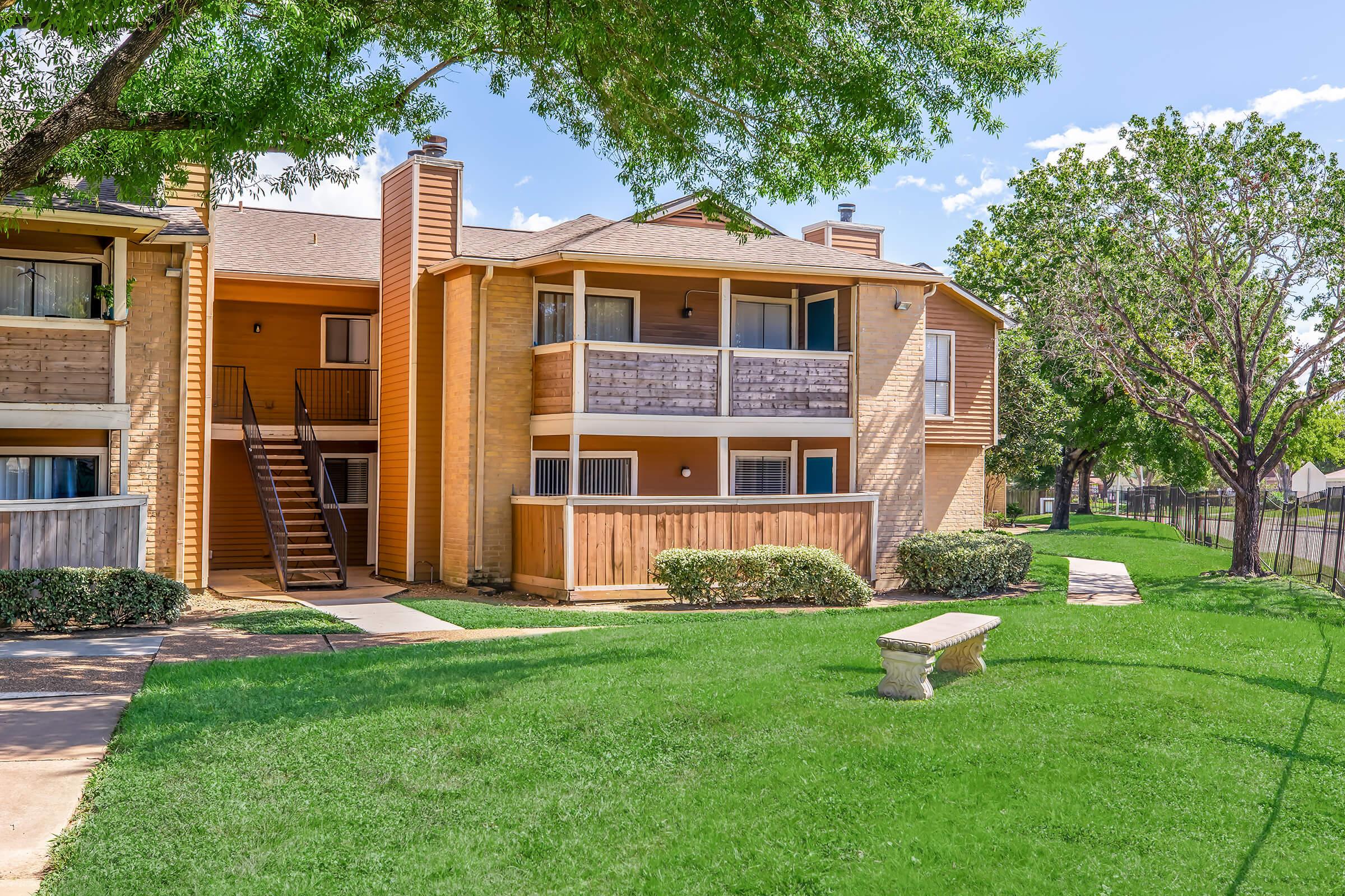 A two-story apartment building featuring a mix of wood and brick exterior, with balconies visible on the upper level. Surrounding green lawn and trees enhance the outdoor space, and a stone bench is positioned along the path leading to the building entrance. Clear blue sky in the background.
