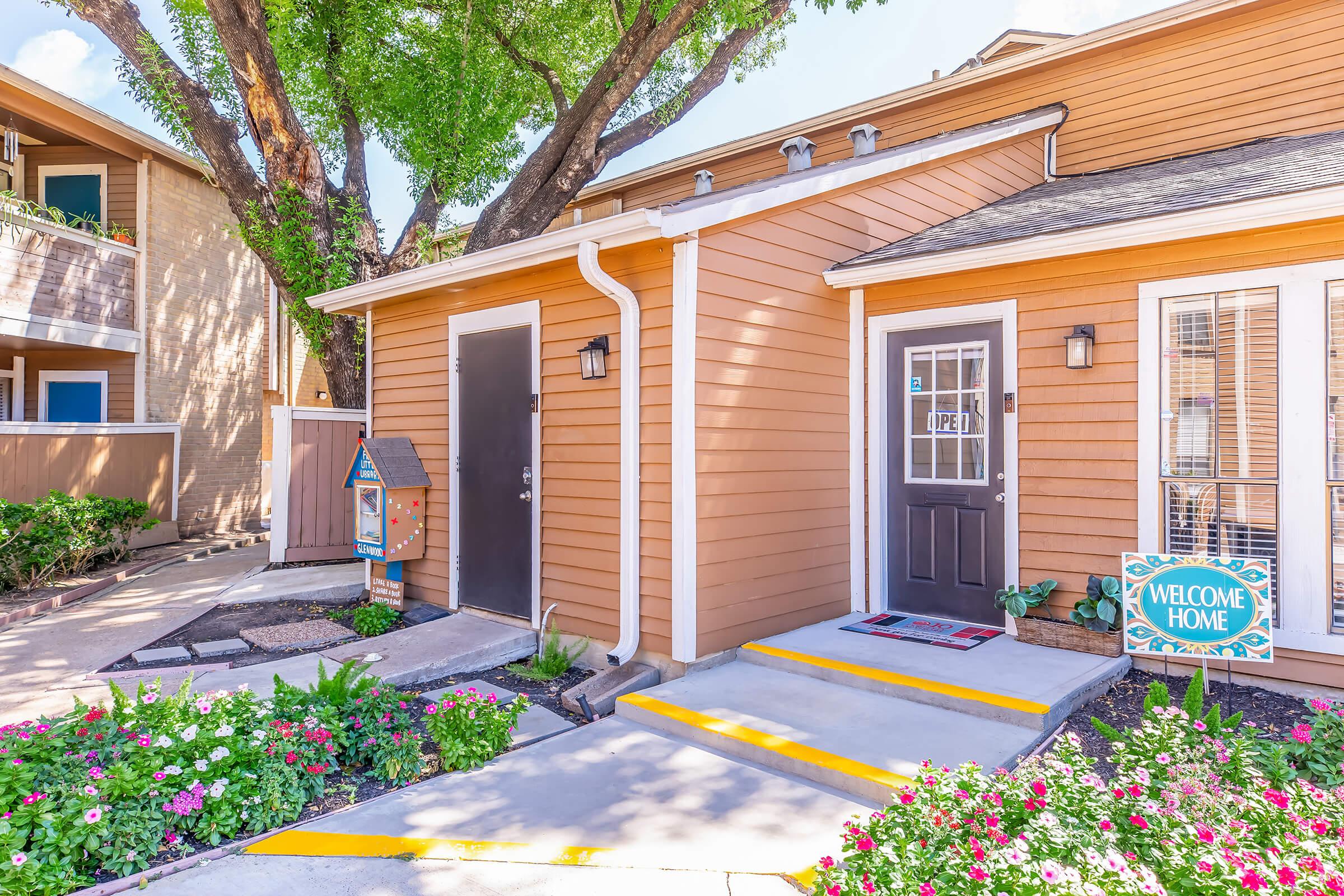 A charming entrance of a residential building featuring two doors, one with a decorative "OPEN" sign. Bright flowers in the foreground, a small mailbox, and a "WELCOME HOME" sign add to the inviting atmosphere. Lush green trees provide shade, creating a cozy, welcoming setting.